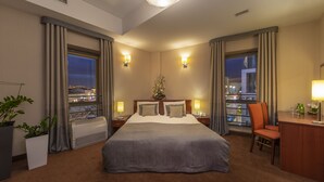 A hotel room with a large bed, two bedside tables with lamps, a desk with a chair, a potted plant, and a view of the city at night. - Hotel Dal Kielce (Kielce)