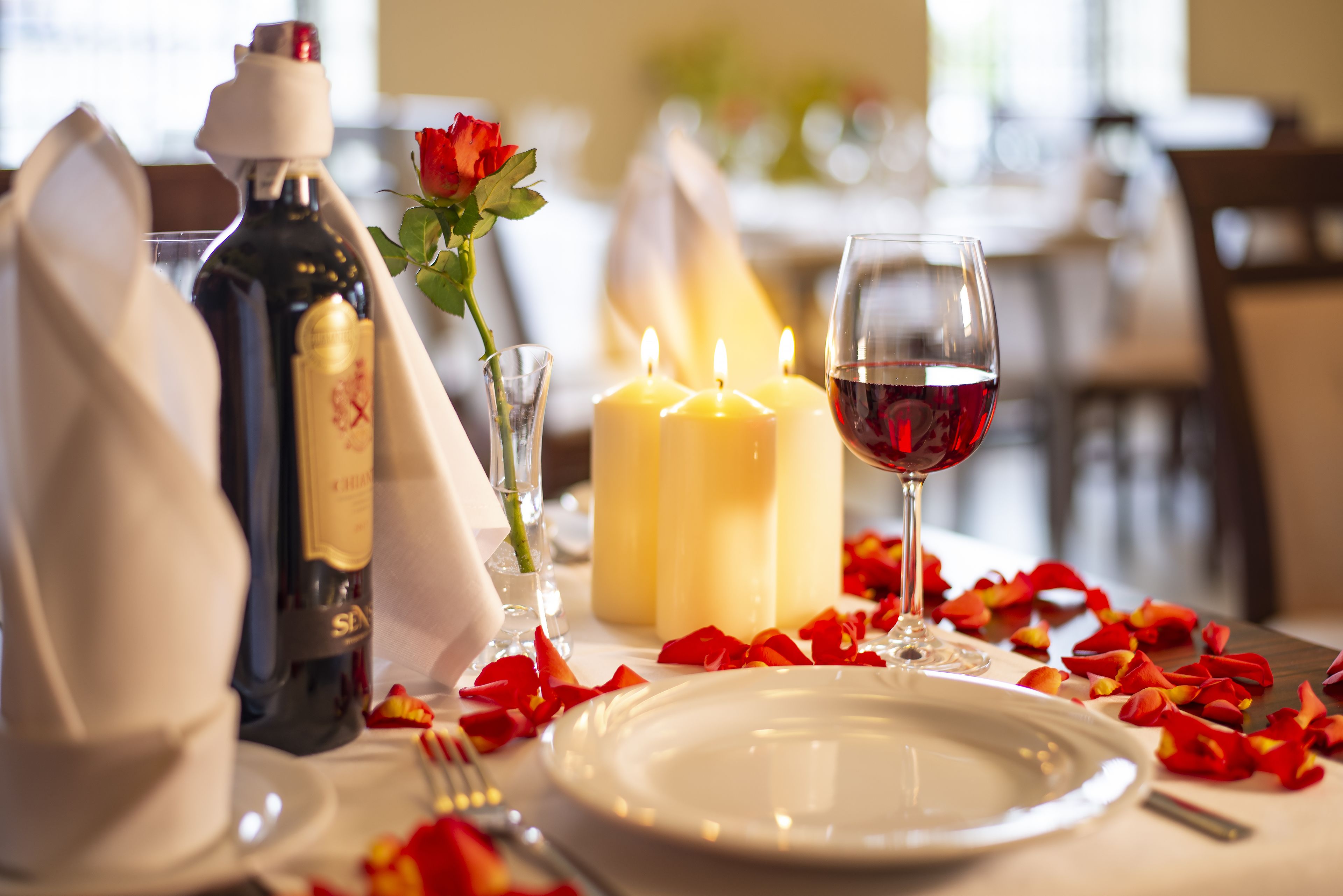 A table set for two with candles, a bottle of wine, a glass of wine, rose petals, and a rose.