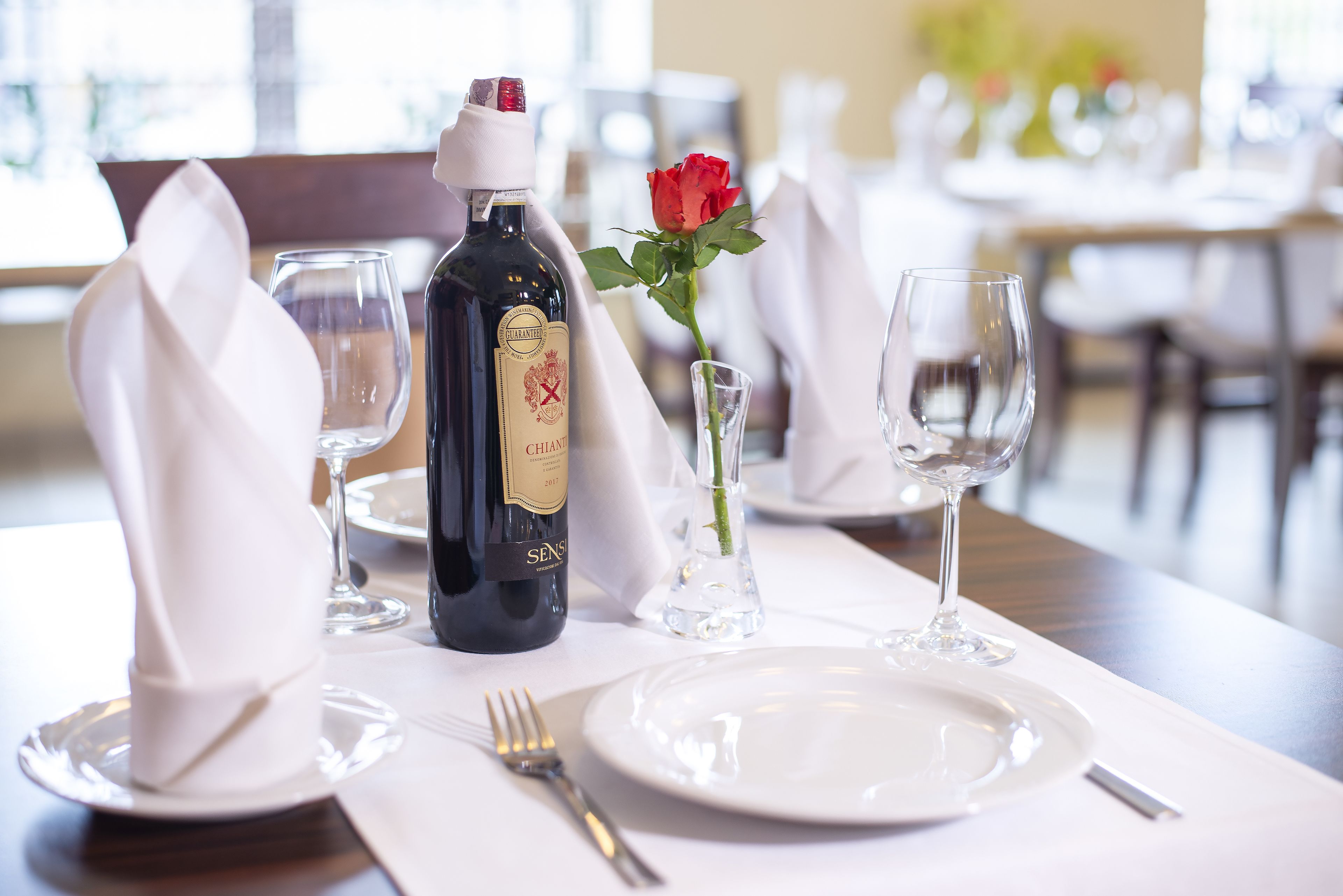 A dining table set with a bottle of wine, a rose, and empty wine glasses.