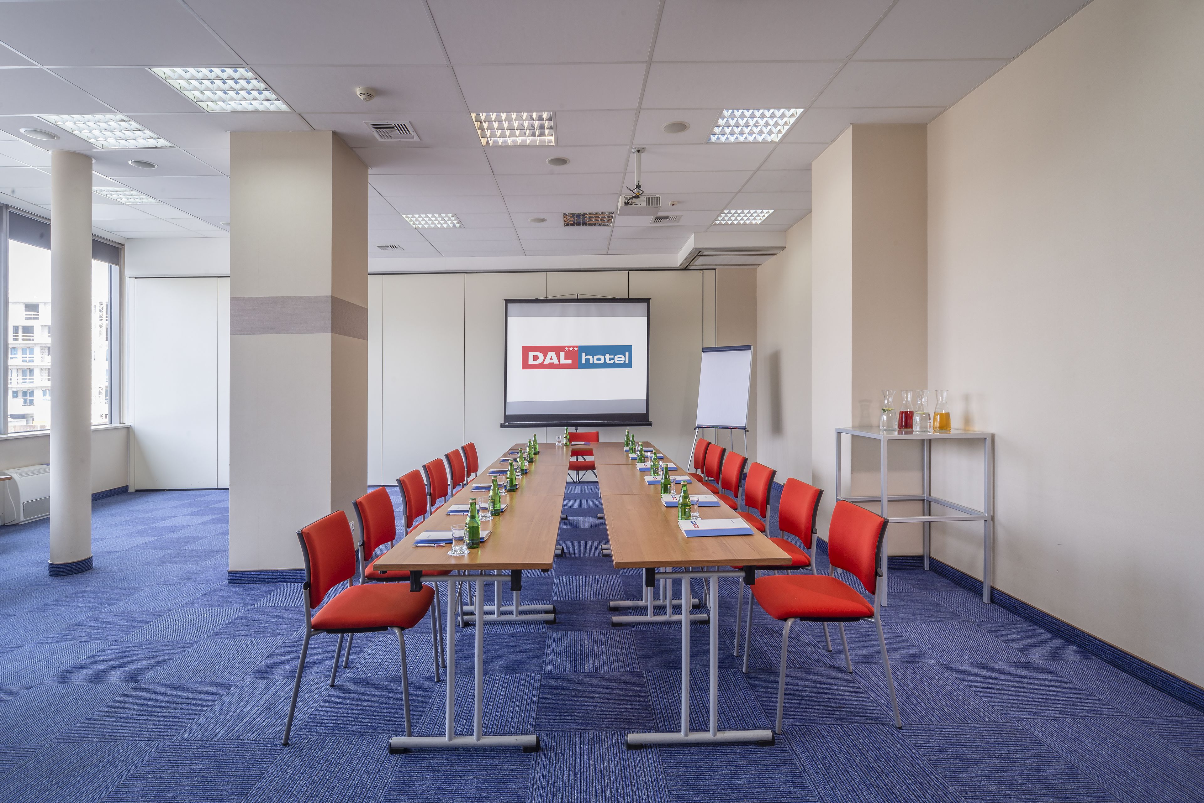 A conference room with a long table, red chairs, and a screen displaying "DAL hotel".