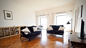 Uma sala de estar ampla com dois sofás pretos, piso de madeira em padrão herringbone, uma estante e uma mesa com computador.