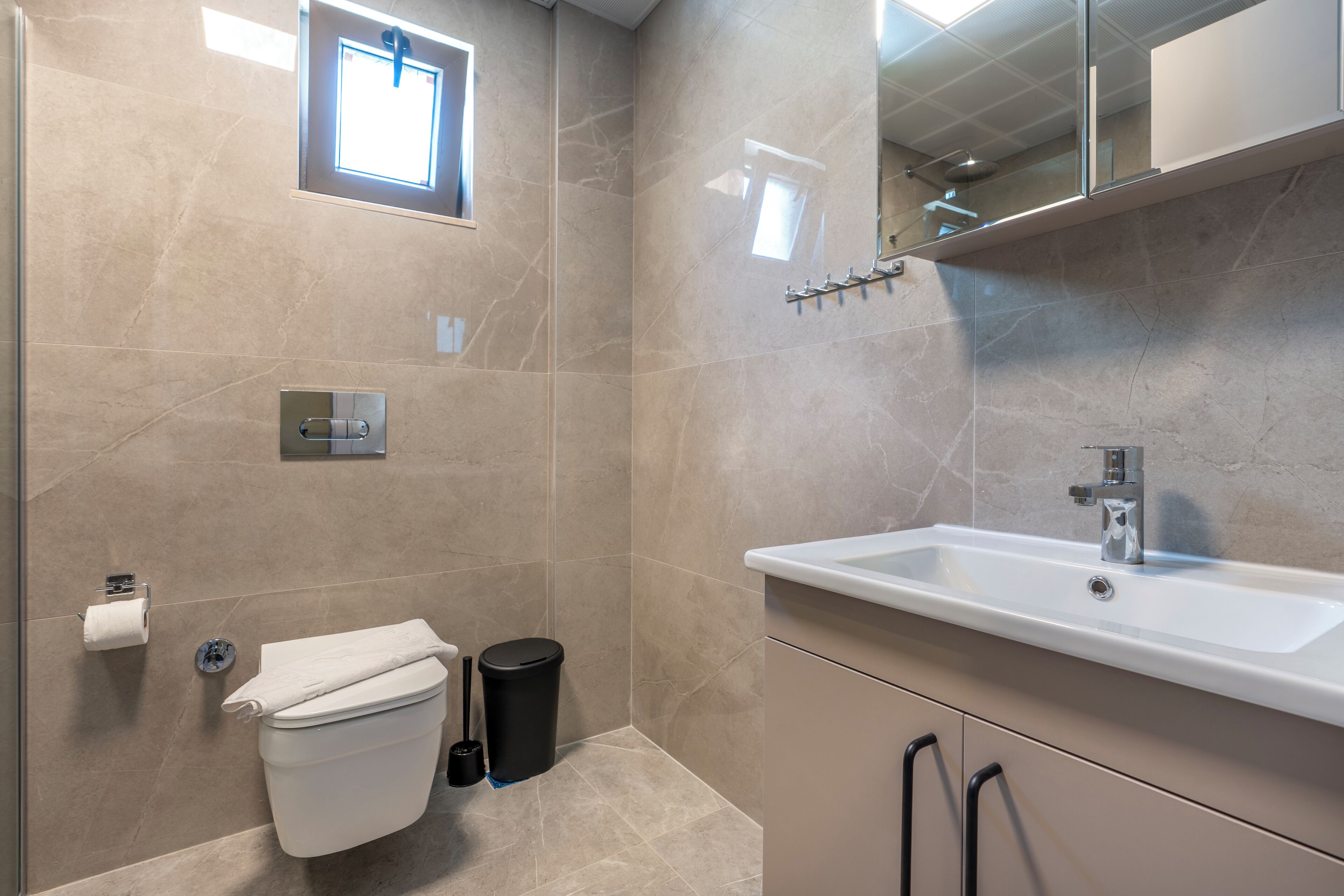 Une salle de bain moderne avec un lavabo blanc, des toilettes, une poubelle et une fenêtre laissant entrer la lumière naturelle.
