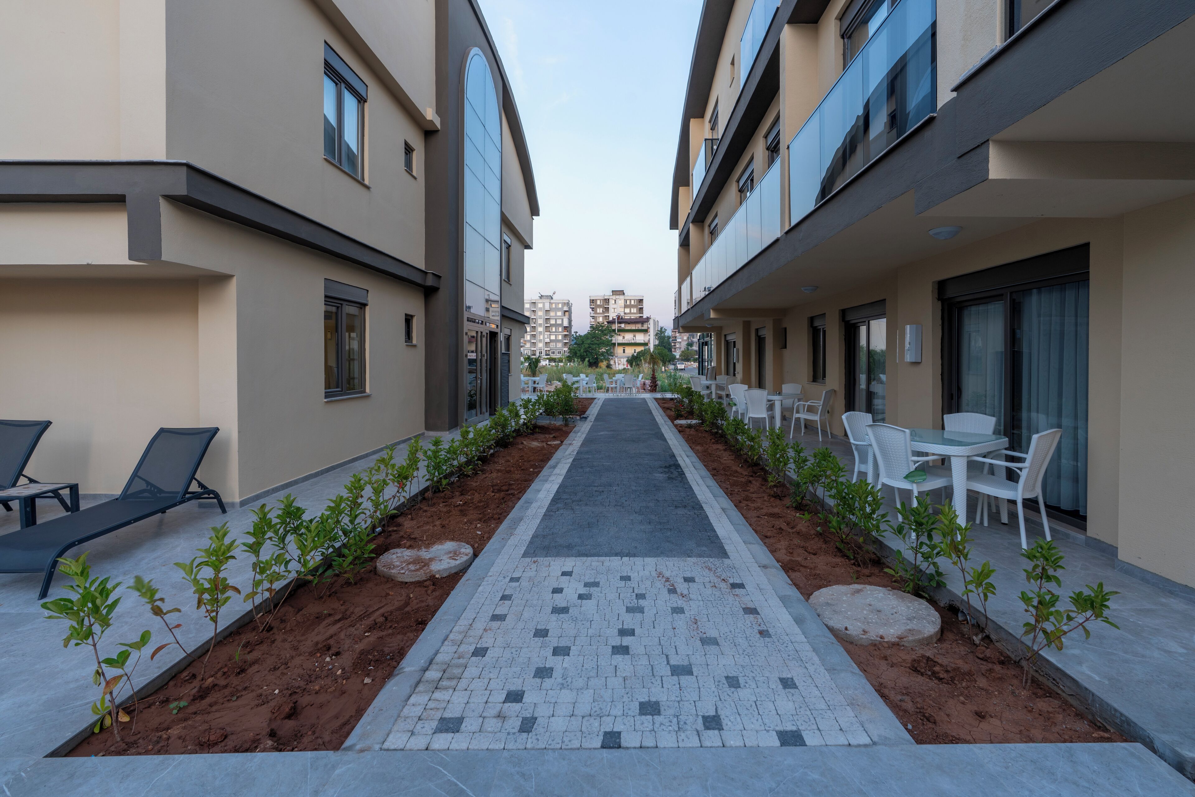 A corridor with a tiled walkway, small plants, and outdoor seating areas with tables and chairs.