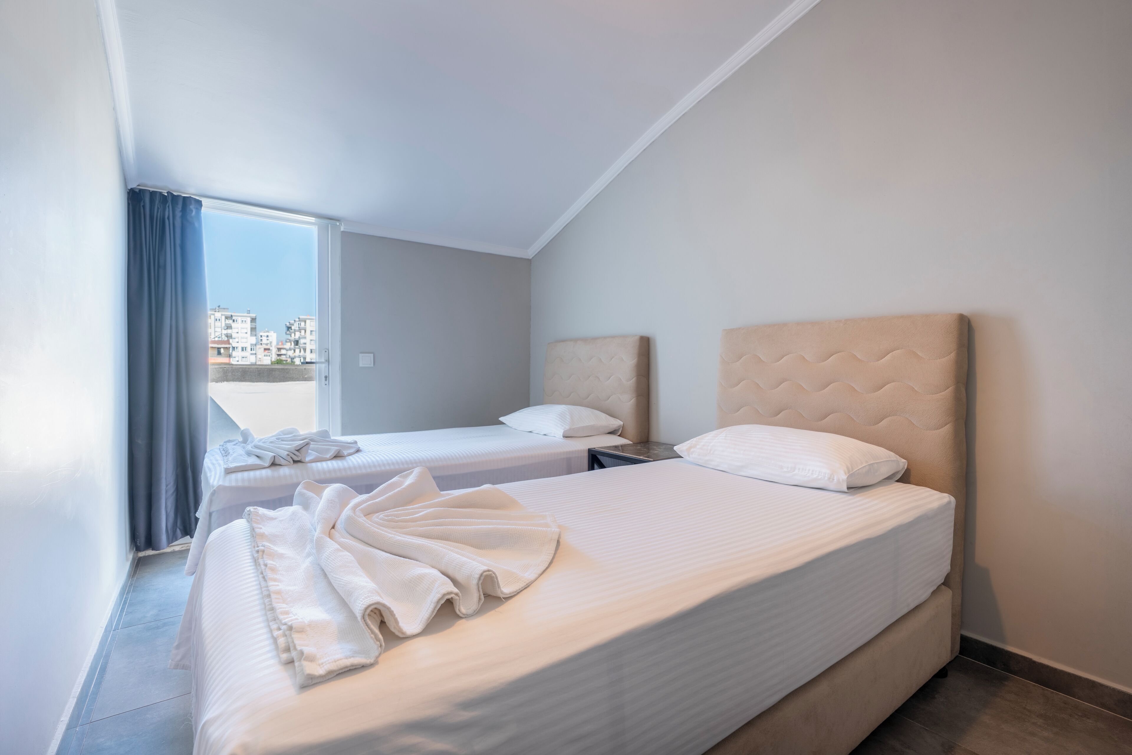 Hotellihuone, jossa on kaksi sänkyä, suuri ikkuna, jossa on verhot, ja näkymä ulkona oleviin rakennuksiin.