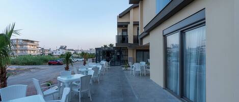 Une terrasse extérieure moderne, avec des tables et des chaises en osier blanc, un ciel dégagé et une vue sur les bâtiments environnants.