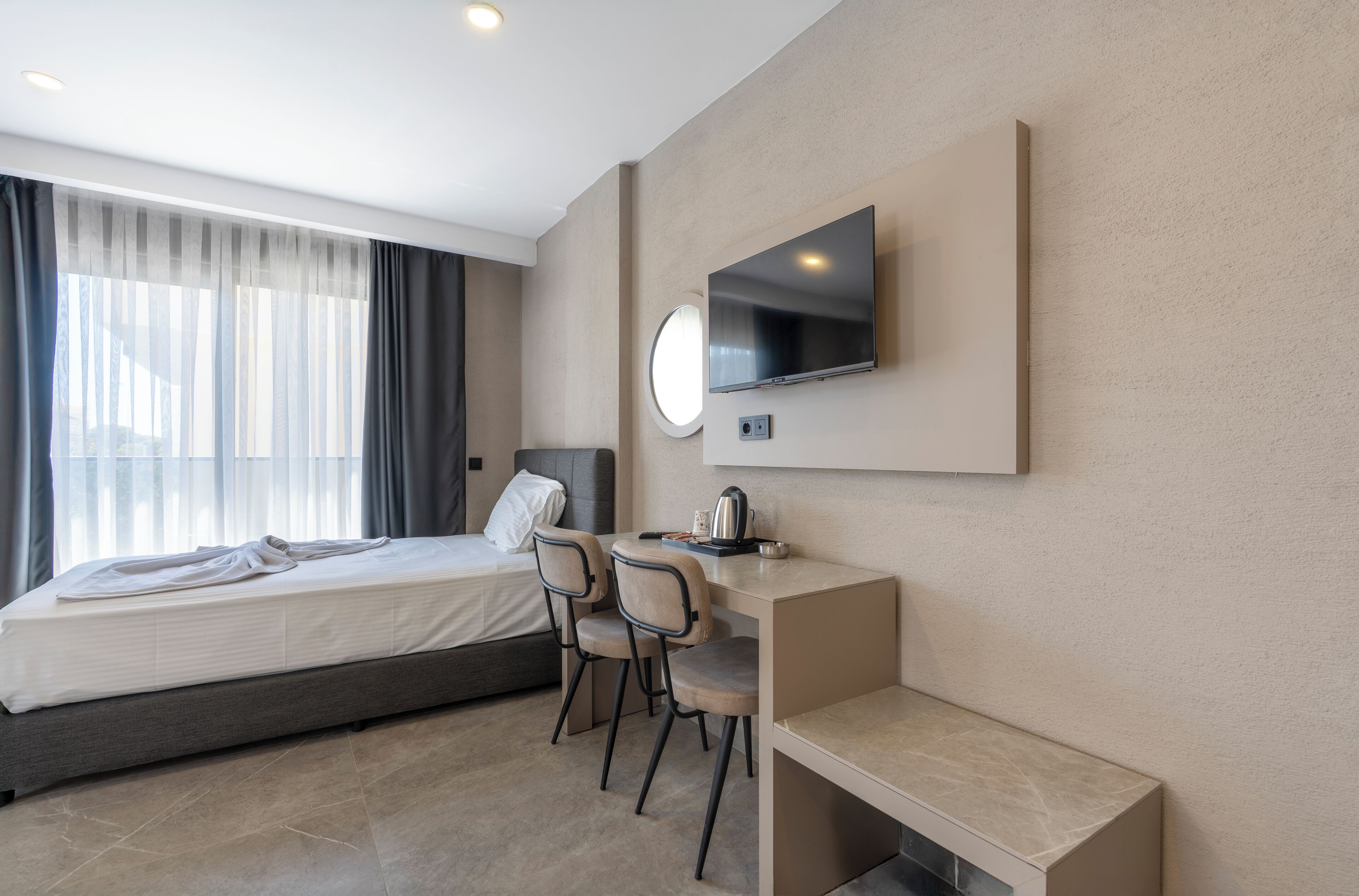 A modern hotel room with a bed, a desk with two chairs, a flat-screen TV mounted on the wall, and a window with curtains.