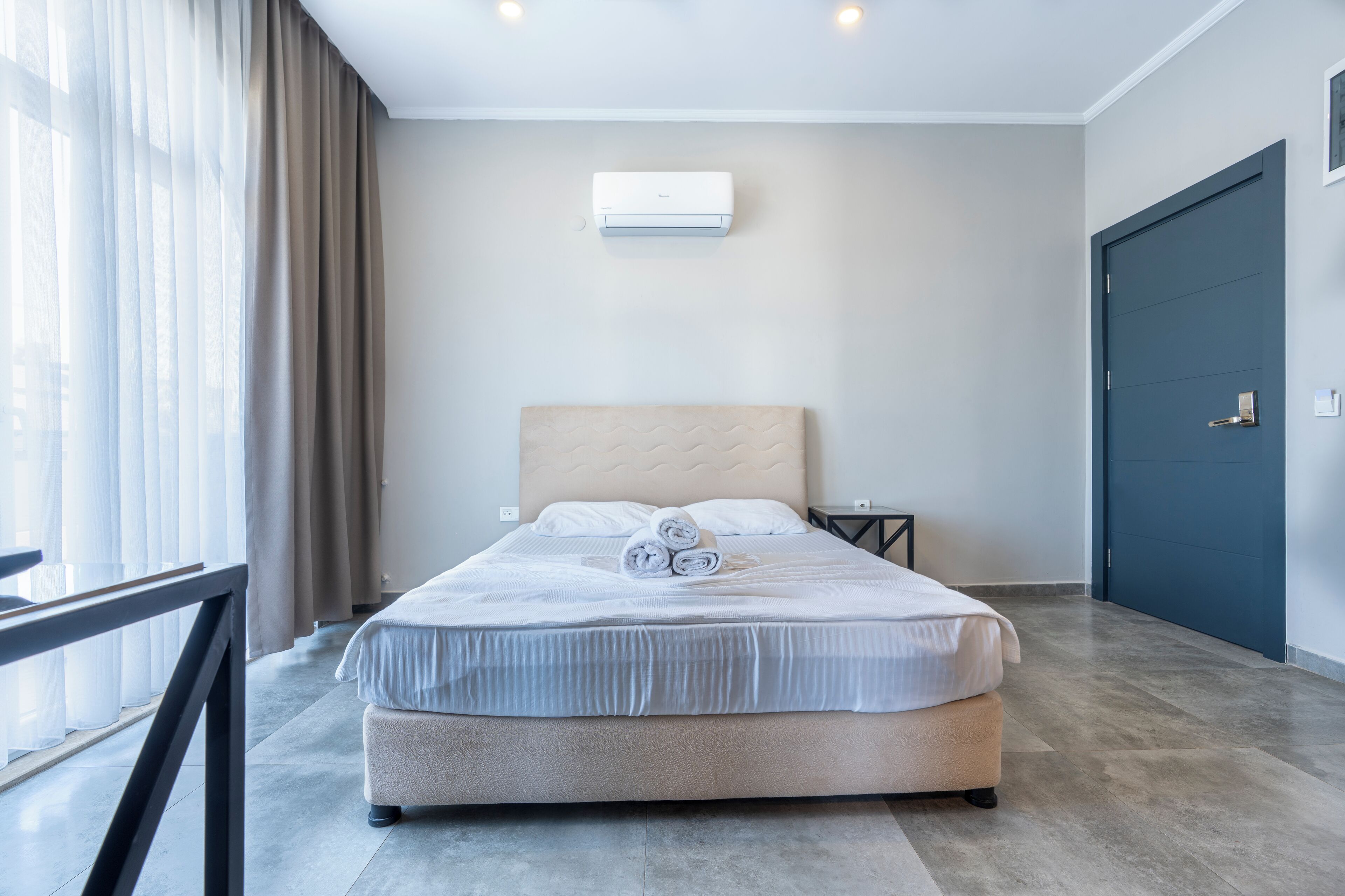 A modern hotel room with a large bed, a headboard, a window with curtains, a wall-mounted air conditioner, and a dark door.