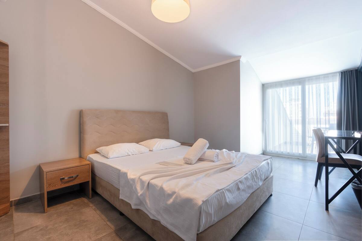 A modern hotel room with a large bed, a bedside table, a chair, and a view of the city through large windows.