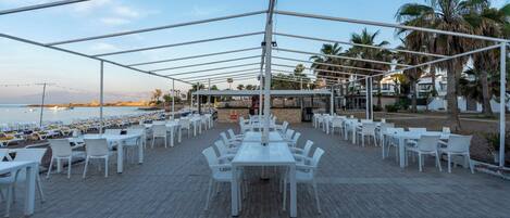 An outdoor dining area with white tables and chairs, a wooden deck, and a view of the sea.