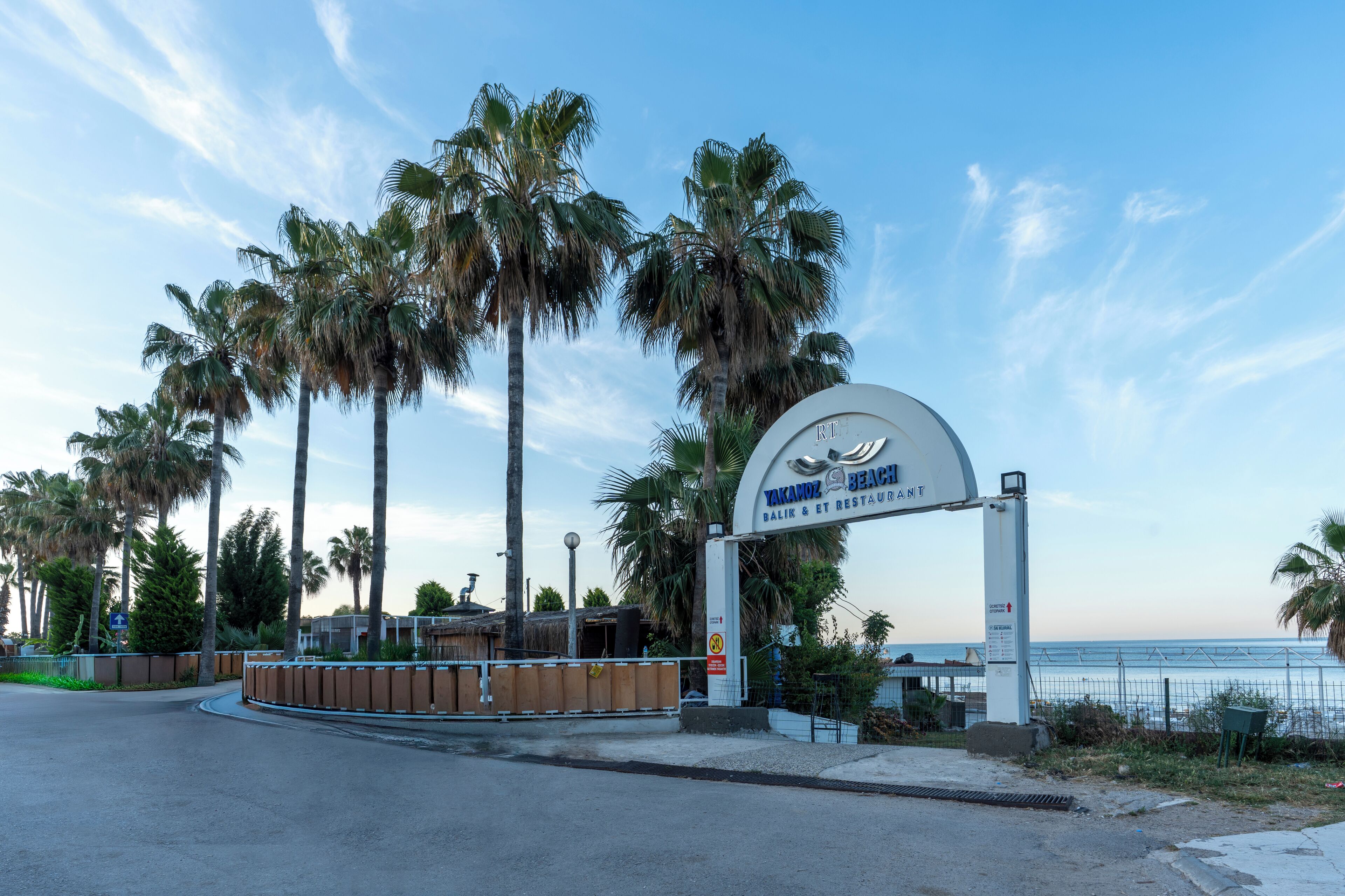 A beach entrance sign for Yoknaptahtzitk Bay Resort & Restaurant with palm trees and a clear sky.