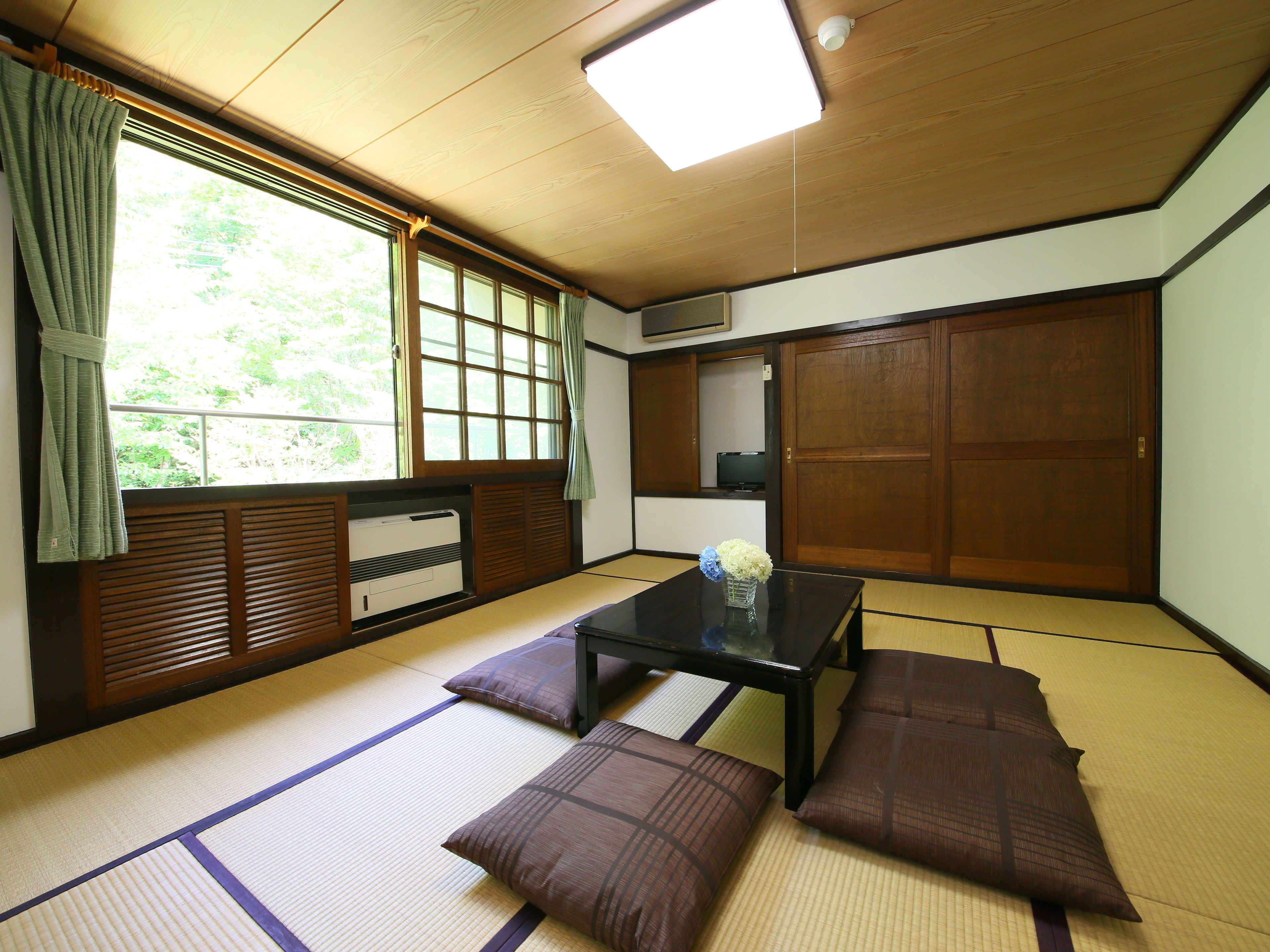 A traditional Japanese room with tatami flooring, a low table, cushions, a window with curtains, and a sliding door.