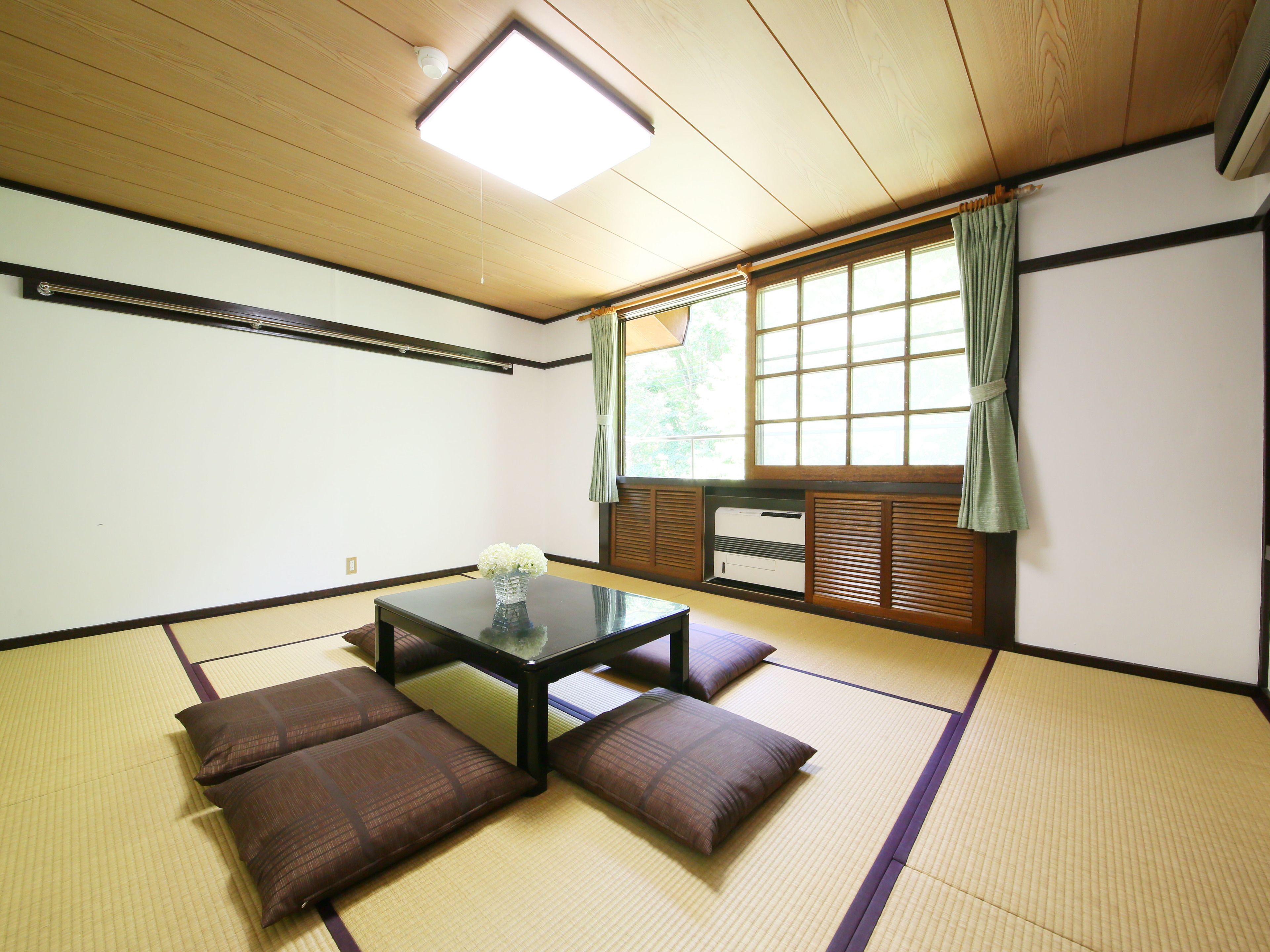 Une pièce avec un sol recouvert de tatamis, une petite table basse, des coussins, un radiateur et une fenêtre avec des rideaux.