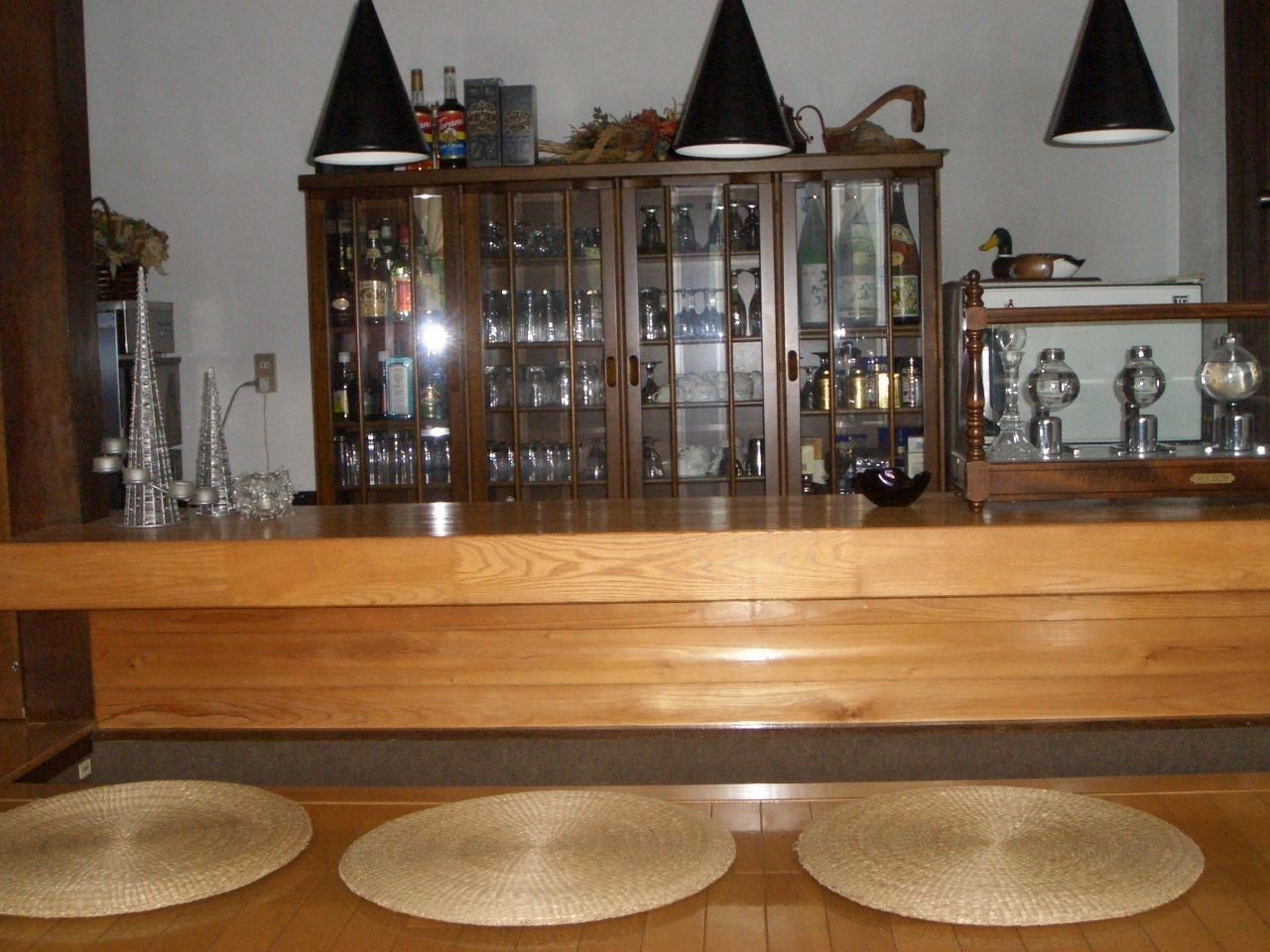 A wooden bar area with a glass cabinet displaying bottles and glasses, two black lamps, and three woven placemats on the floor.