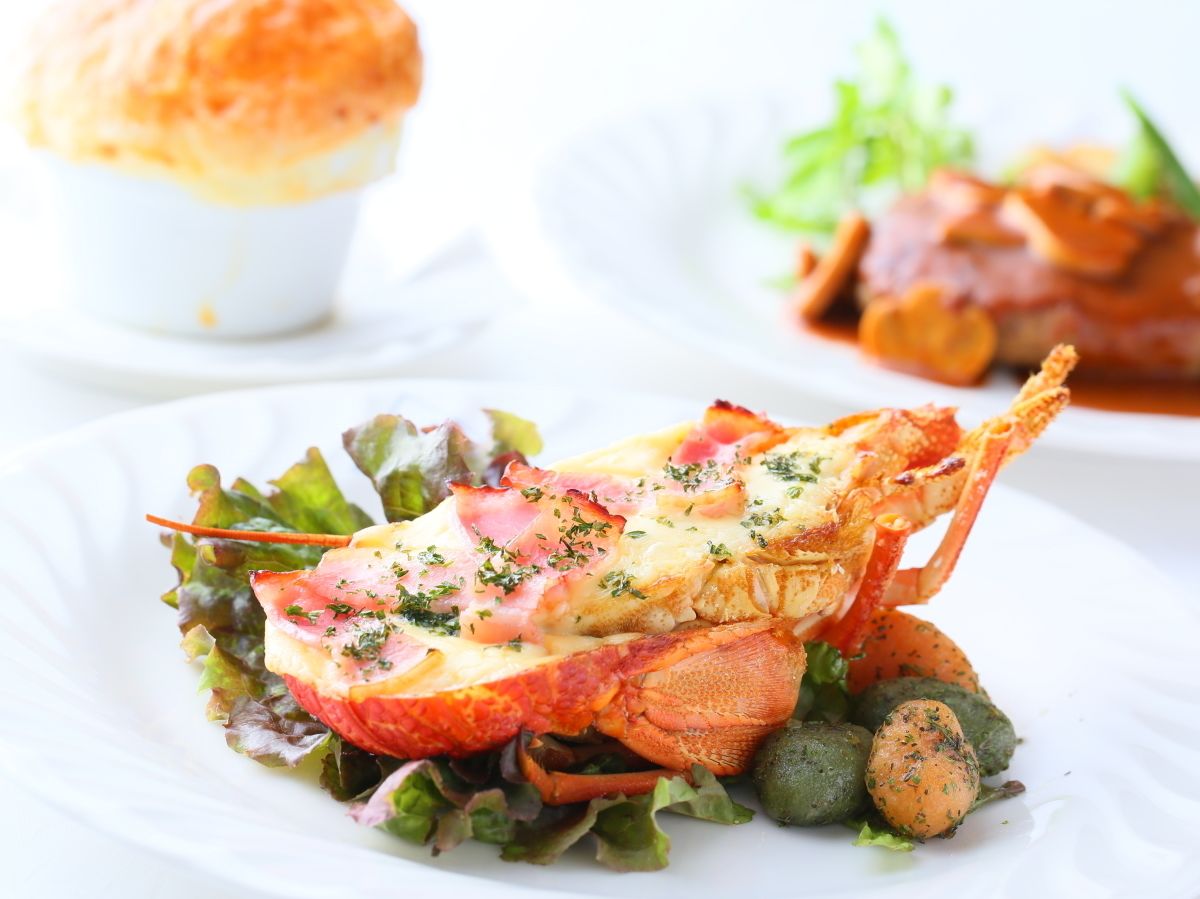 A lobster tail with melted cheese and herbs on a bed of greens, served on a white plate.