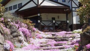 A building with a stone staircase leading to a covered entrance, surrounded by a variety of flowering plants. - Chalet La Neige (Minakami)