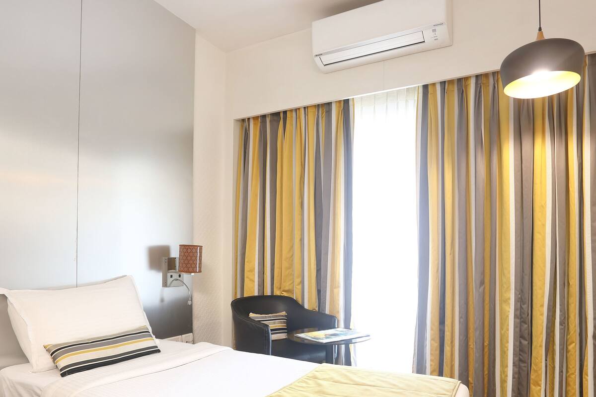 A hotel room with a bed, a desk, a chair, a ceiling fan, an air conditioner, and curtains.