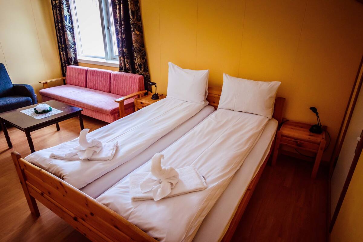 Cama bem arrumada com lençóis e toalhas brancos, mesa de cabeceira de madeira, pequena mesa com abajur, sofá rosa e cadeira azul em um quarto com piso de madeira e paredes amarelas.