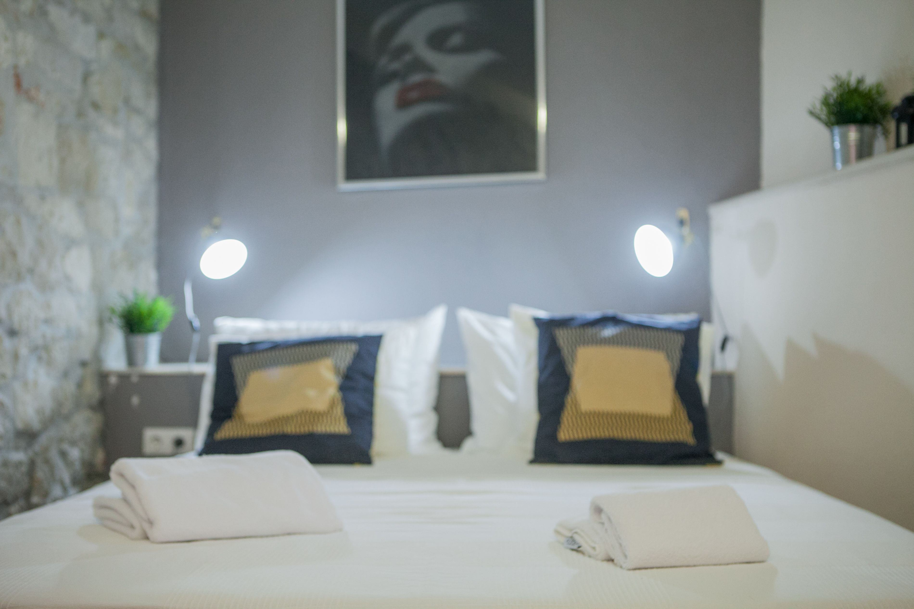 A neatly made bed with white linens and two decorative pillows, a bedside table with a small plant, and a wall-mounted light fixture.