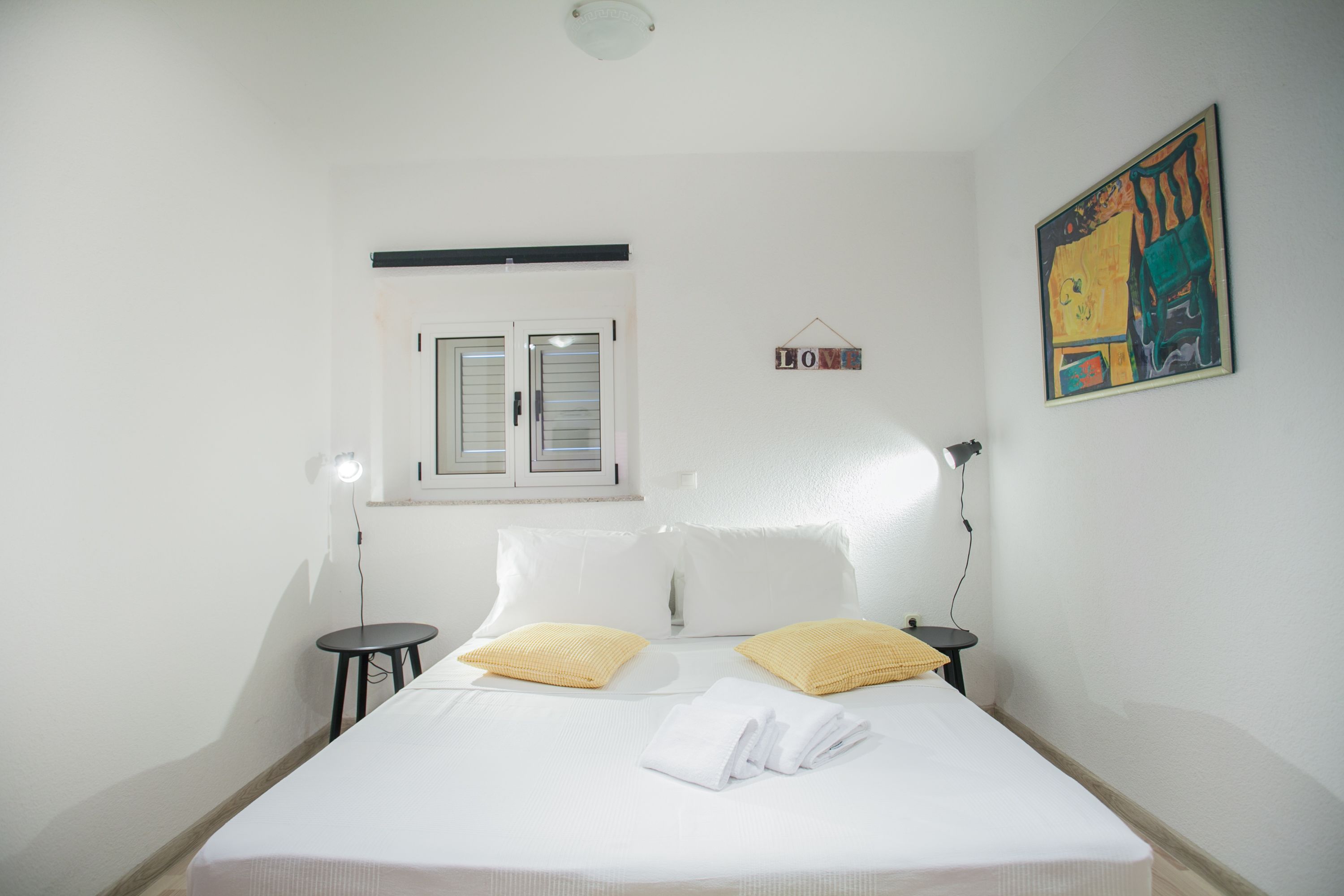 A neatly made bed with white linens and two yellow pillows, a small bedside table with a lamp, a window with shutters, and a wall-mounted artwork.