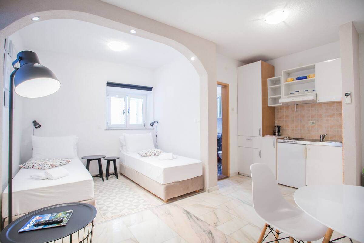 Une chambre d’hôtel de taille réduite, comprenant un lit, une petite table, une chaise, un bureau et un coin cuisine.