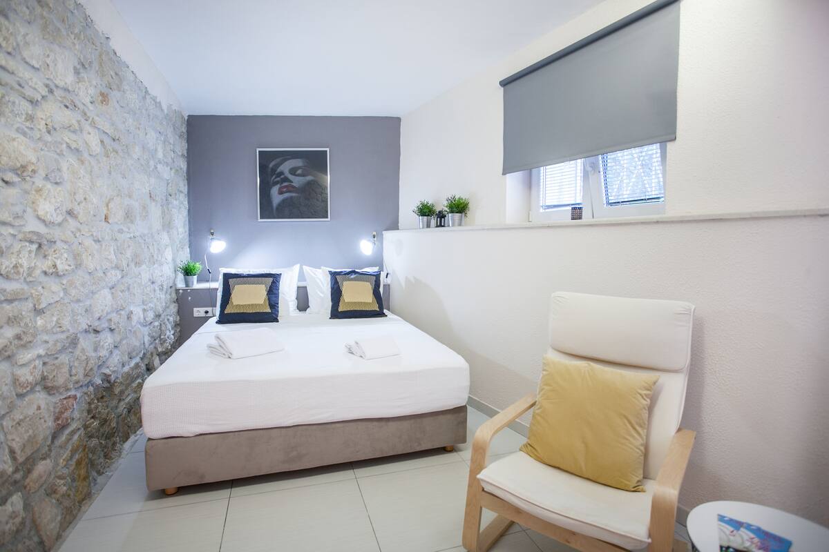 Une chambre avec un mur en pierre, un lit avec une literie blanche, deux tables de chevet avec des lampes, une fenêtre avec des stores et une chaise avec un coussin.