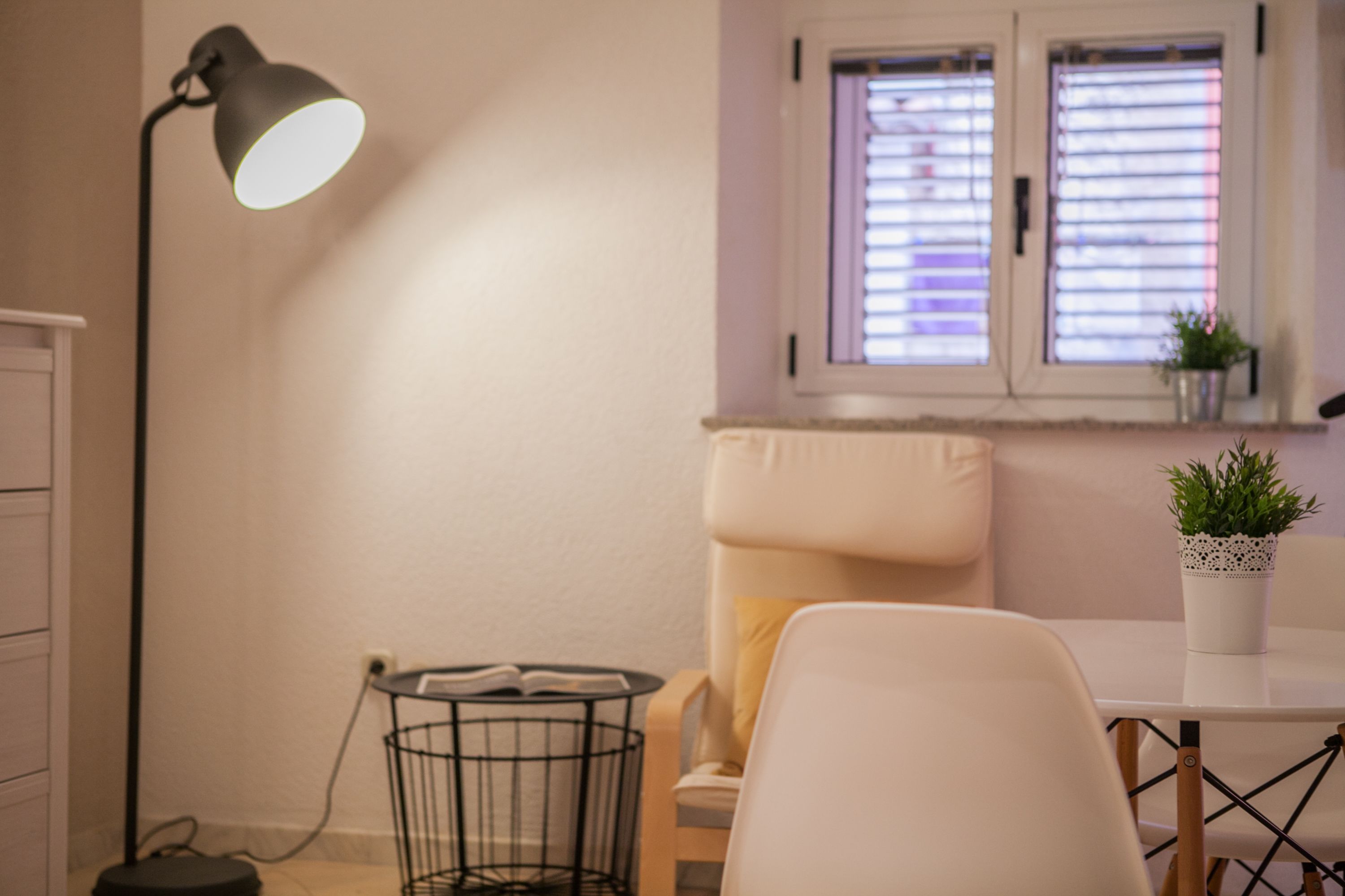 Ein Zimmer mit weißem Sessel, einem kleinen Tisch mit Topfpflanze, einer Stehlampe und einem Fenster mit Jalousien.