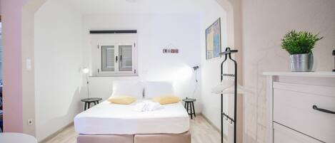 Ein Schlafzimmer mit Bett, Nachttischen, einem Schminktisch, einer Zimmerpflanze und einem Fenster mit Fensterläden.