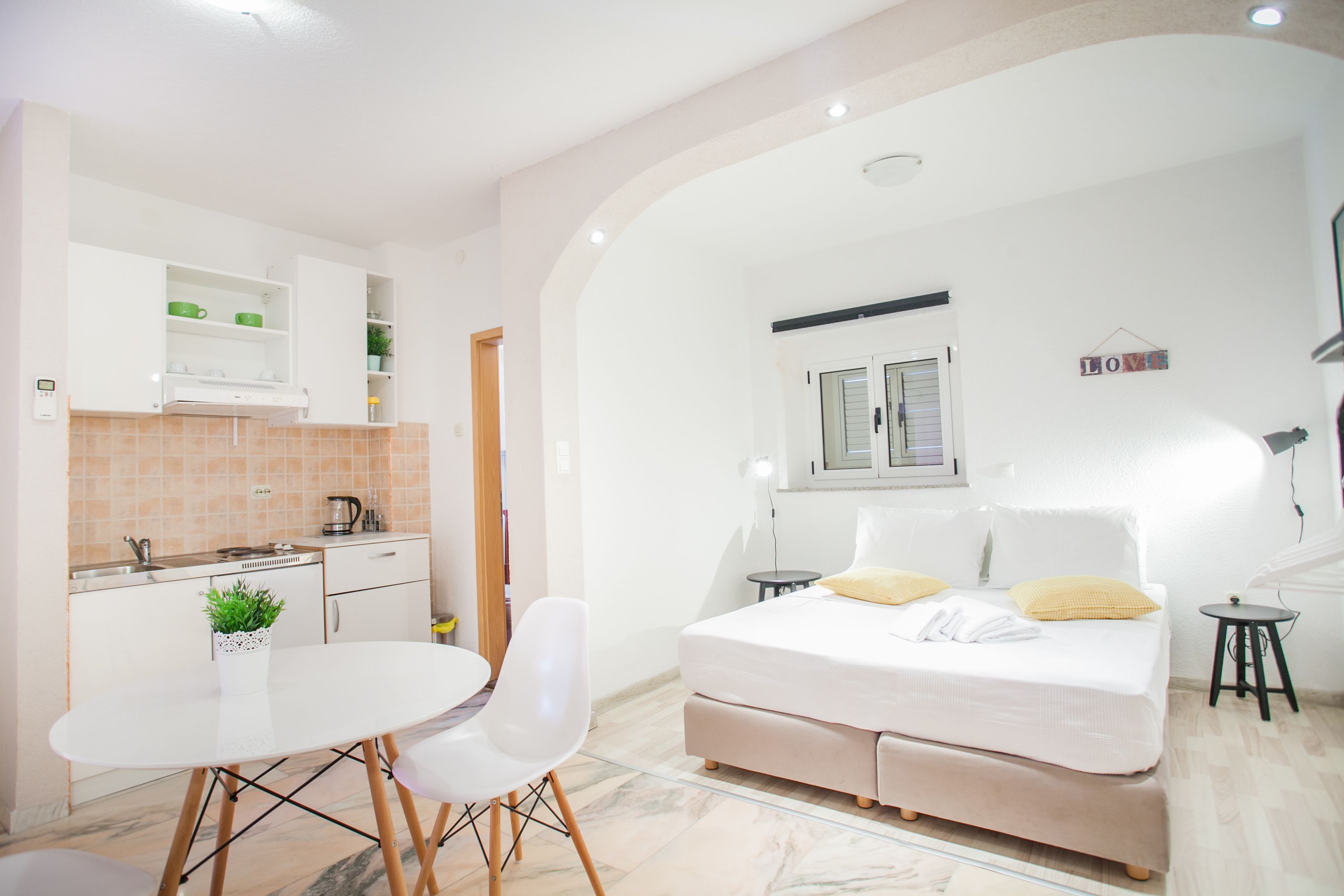 Una habitación compacta con kitchenette, una cama con dos almohadas, una mesita pequeña y una silla.