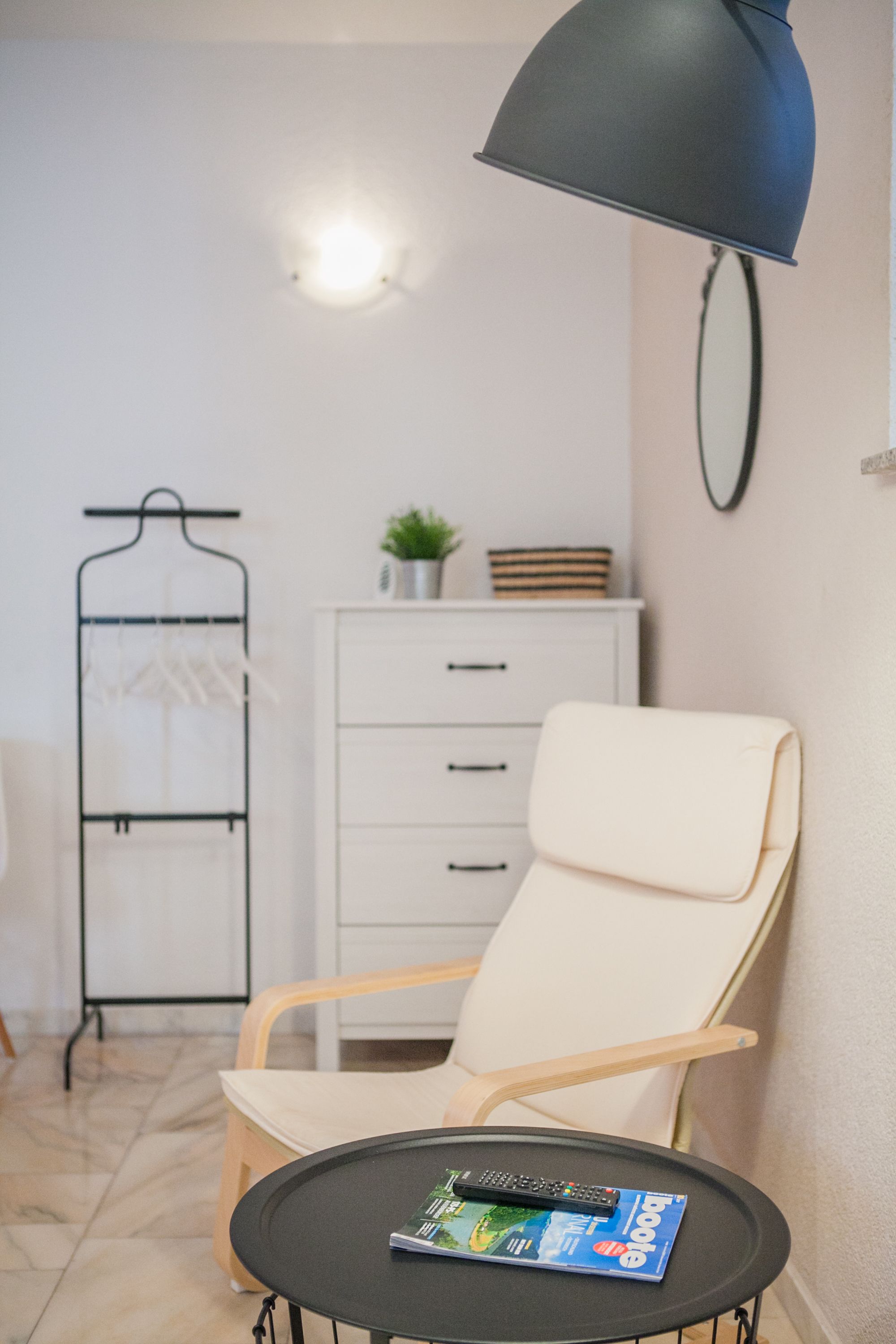 Een moderne kamer met een witte stoel, een klein tafeltje met een afstandsbediening en een tijdschrift, een metalen kapstok met kleding, een witte kast met lades, en een wandlamp.