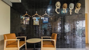 A cafe with two wooden chairs and a small round table, a black tiled wall, and a sign that reads "Cafe" with hats hanging above. - Lazy Sunday Hostel (Bangkok)