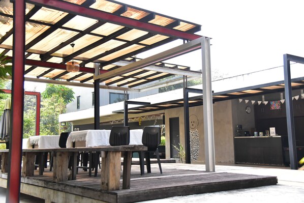 Un patio al aire libre con mesa y sillas de madera, una zona de estar cubierta y un edificio moderno al fondo.