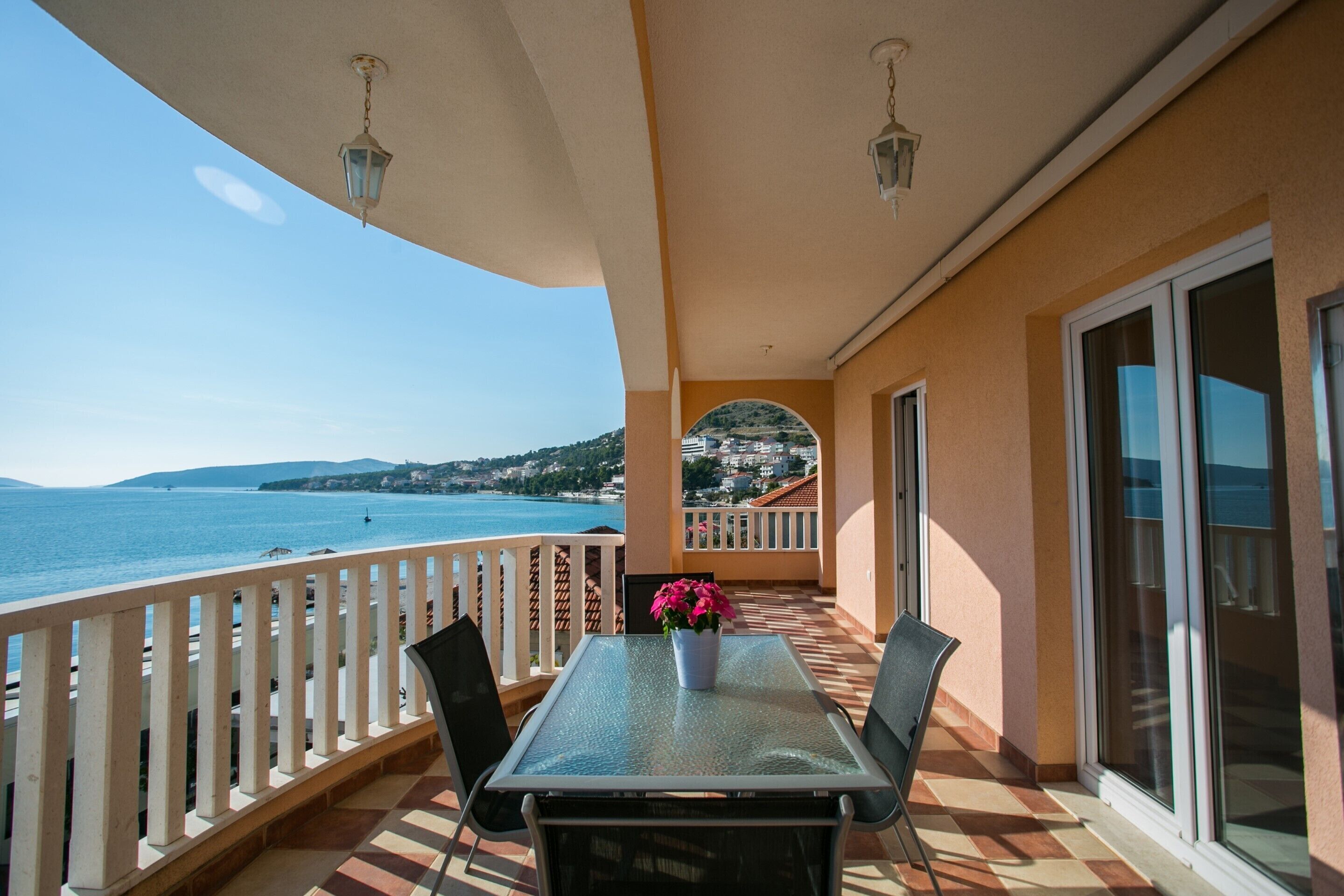 A balcony with a table and chairs, a potted plant, and a view of the sea and distant hills.