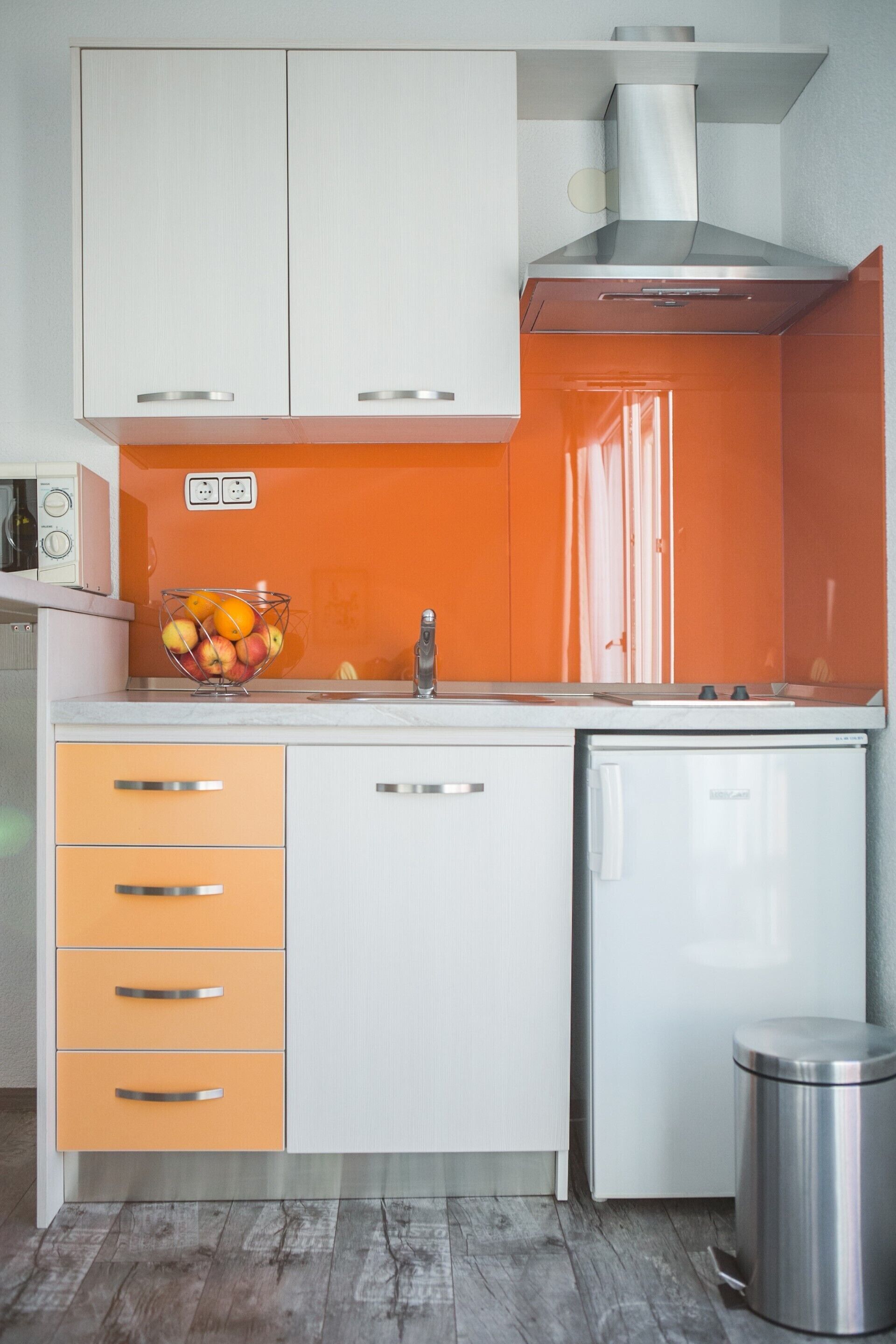 Een kleine keuken met witte kastjes, een oranje spatwand, een roestvrijstalen spoelbak en een kleine koelkast.