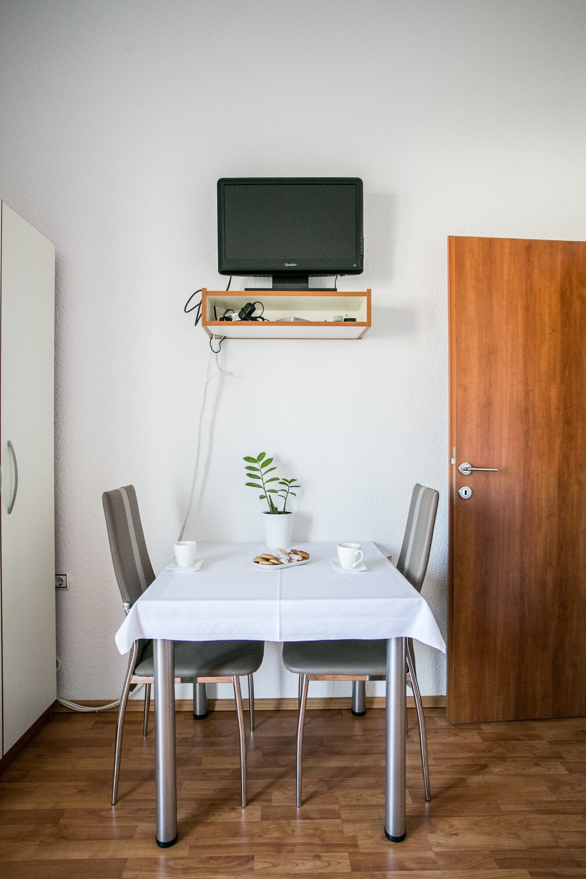 Et spiseområde med et bord dækket til to personer, en potteplante og et ophængt fjernsyn over bordet.