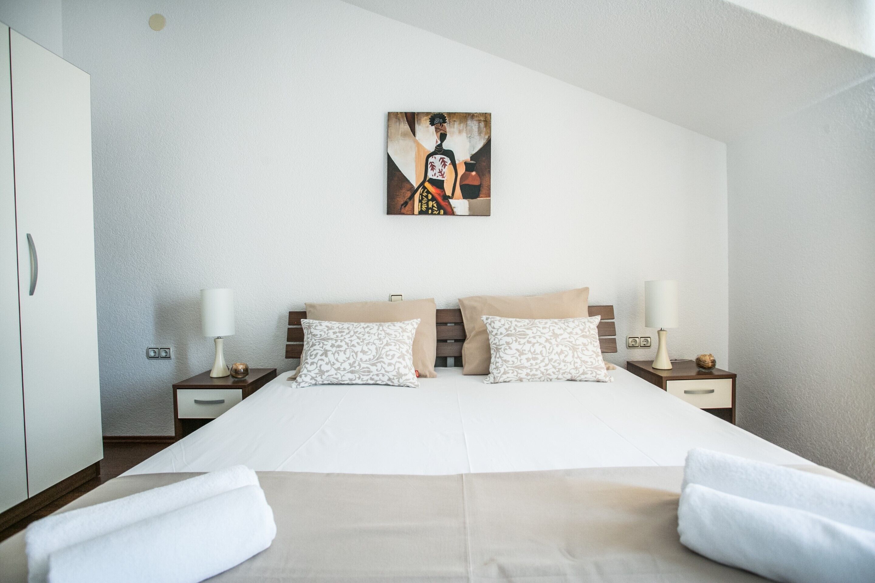 A hotel room with a large bed, two bedside tables, a painting on the wall, and a wardrobe.