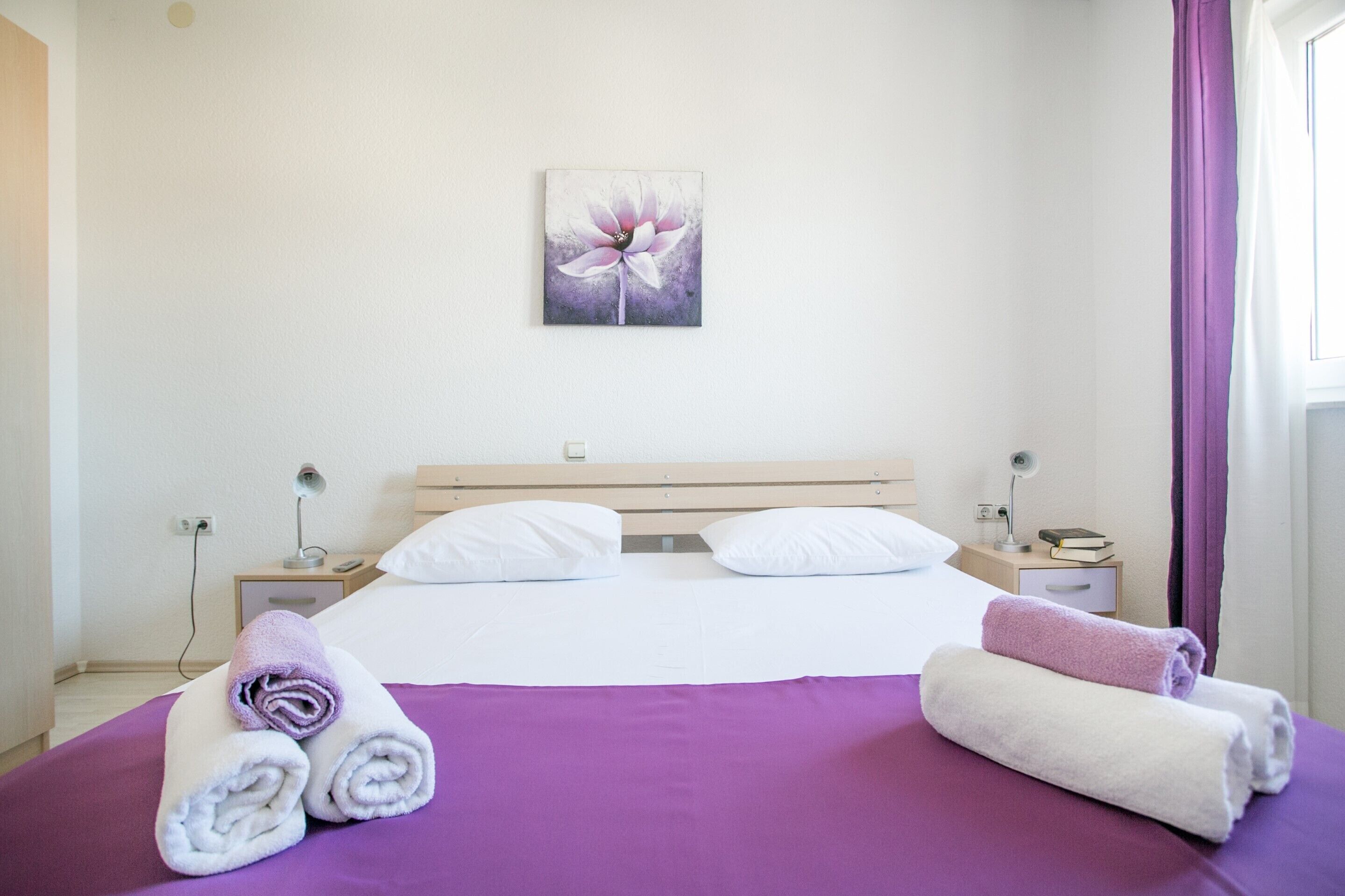A hotel room with a bed, two white pillows, a purple bedspread, a painting of a flower on the wall, and two nightstands with lamps and books.