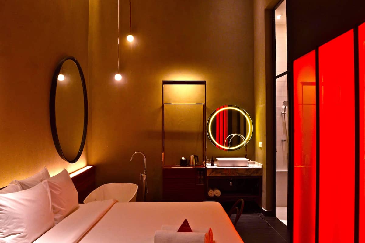Een hotelkamer met een bed, nachtkastje, spiegel, badkamer met wastafel en spiegel, en een rode accentmuur.