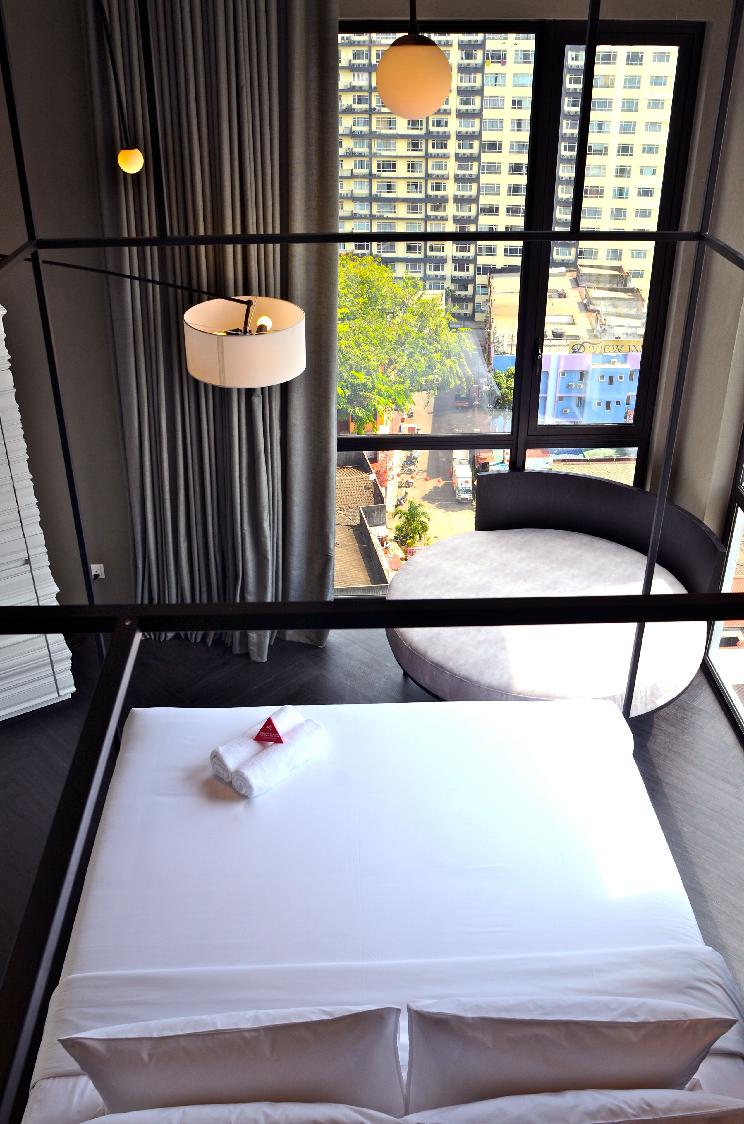 A hotel room with a large window, a bed with white linens, a round seating area, and a hanging light fixture.