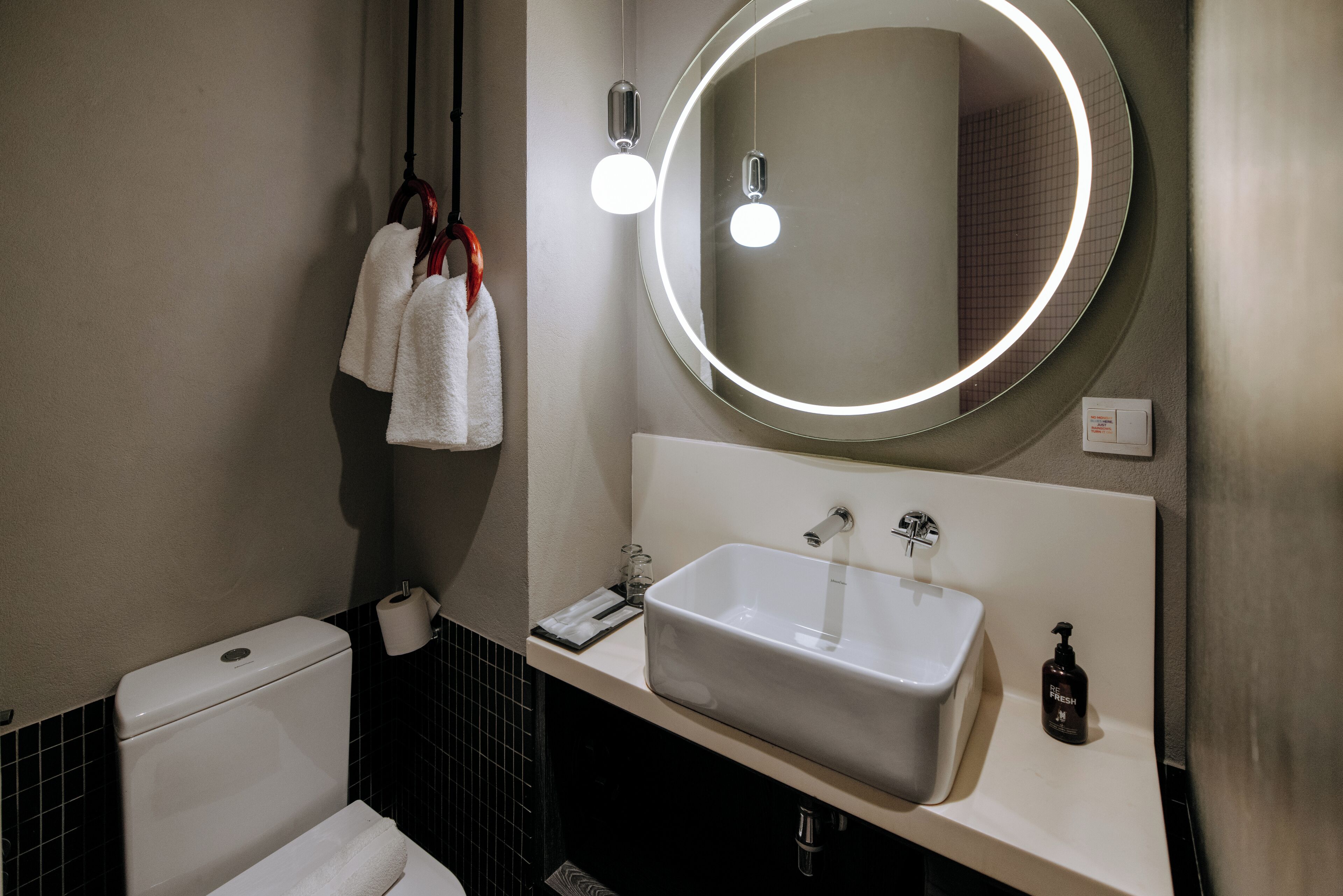 A bathroom with a white sink, a round mirror with lights, a towel rack with towels, and a toilet.