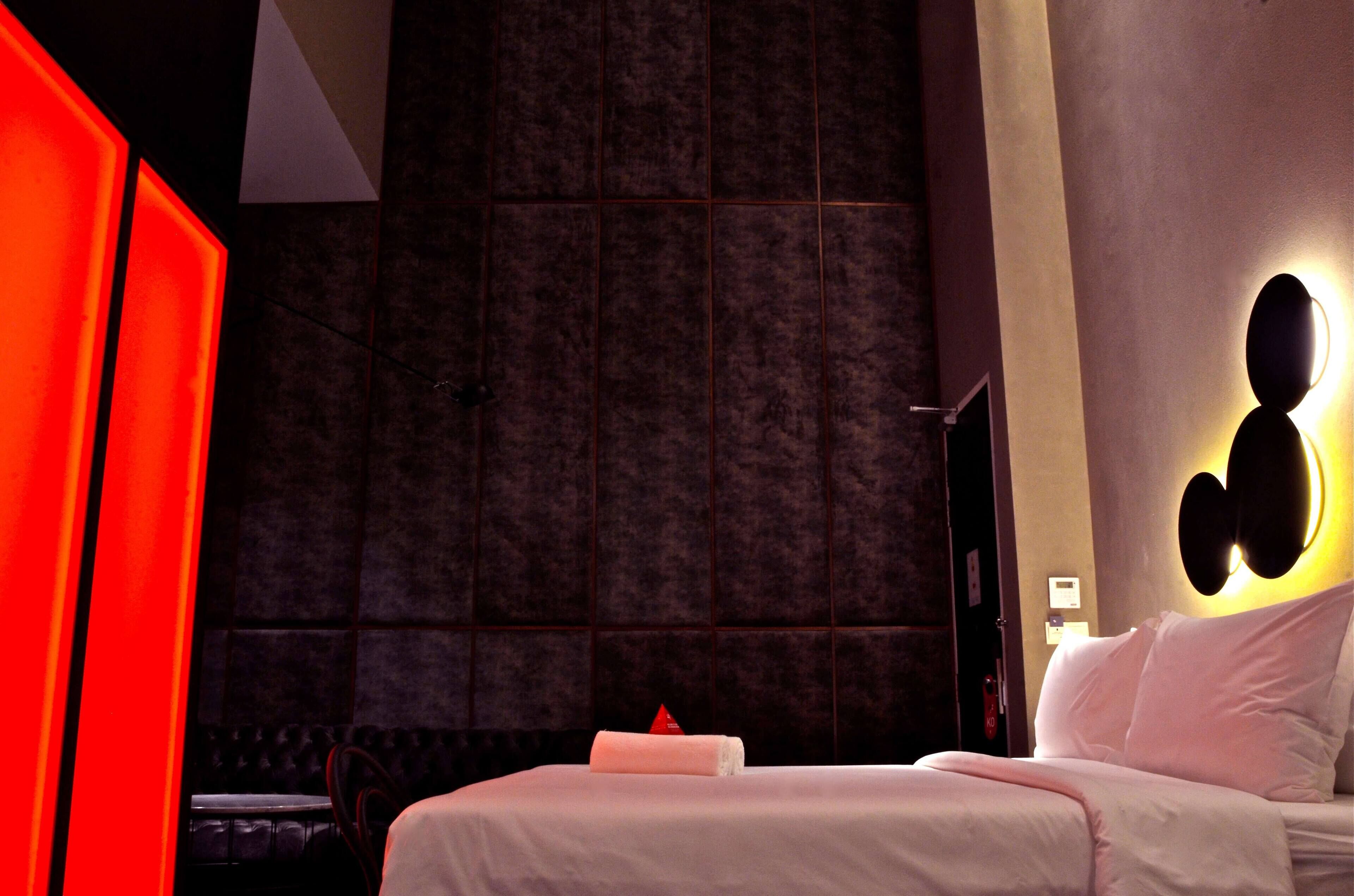 A modern hotel room with a large bed, a red accent wall, and a unique wall-mounted light fixture.