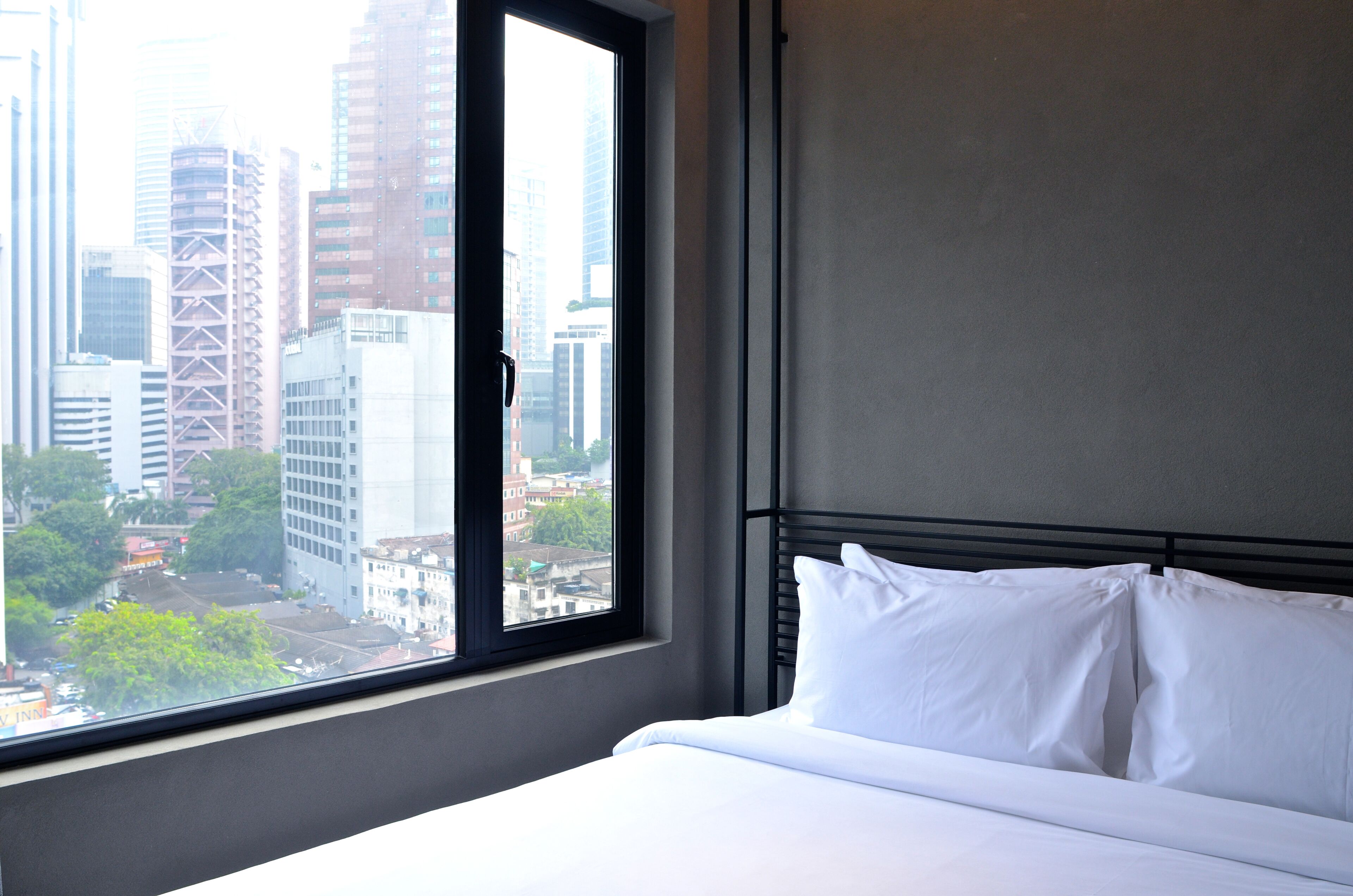 A hotel room with a large window offering a city view, a neatly made bed, and a simple headboard.