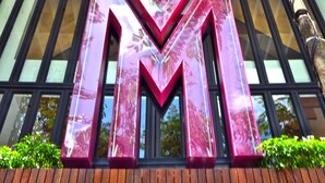 Un grand panneau en forme de « M », brillant et bien visible, se trouve devant un bâtiment à l’architecture moderne, doté de grandes fenêtres, et un arbre est visible au premier plan.