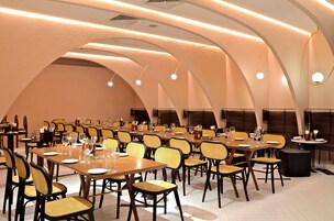 A spacious dining area with multiple tables and chairs, featuring a modern design with curved ceiling lines and pendant lighting.