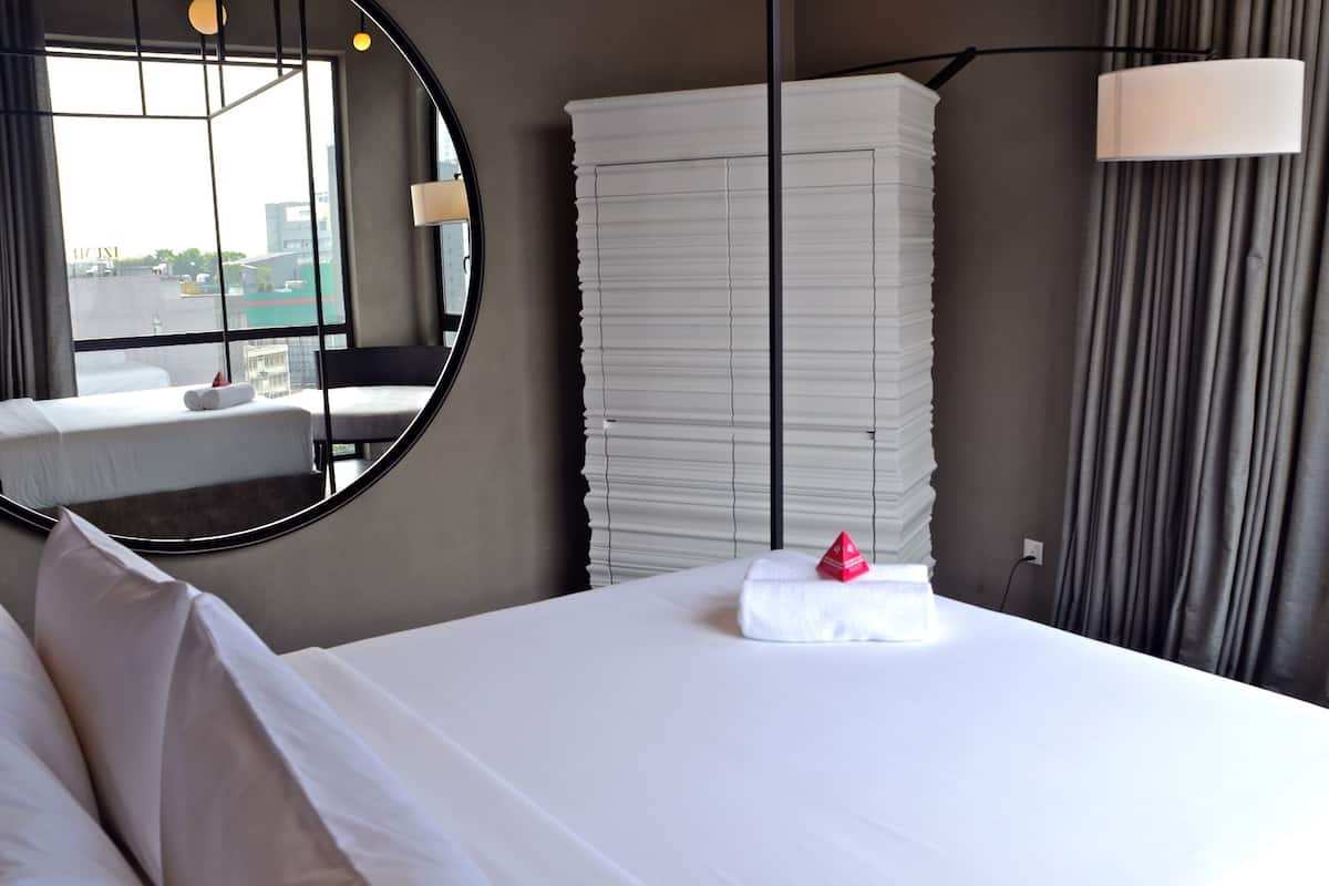 Een hotelkamer met een groot bed, een ronde spiegel, een raam met uitzicht op gebouwen, een stapel witte gordijnen en een lamp.