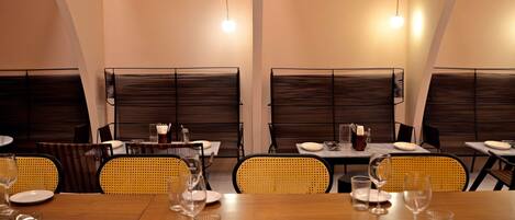 A dining area with tables set for four, each with plates, glasses, and cutlery. The room has a modern design with dark paneling on the walls and yellow woven chairs.