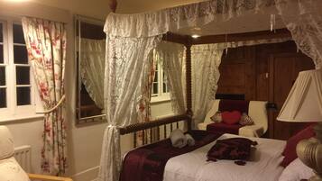 A four-poster canopy bed with white linens, a wooden headboard, and a red throw blanket. There is a window with floral curtains, a wooden chair with a heart pillow, and a bedside table with a lamp.