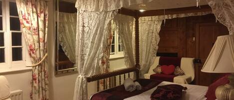 A four-poster canopy bed with white linens, a wooden headboard, and a red throw blanket. There is a window with floral curtains, a wooden chair with a heart pillow, and a bedside table with a lamp.