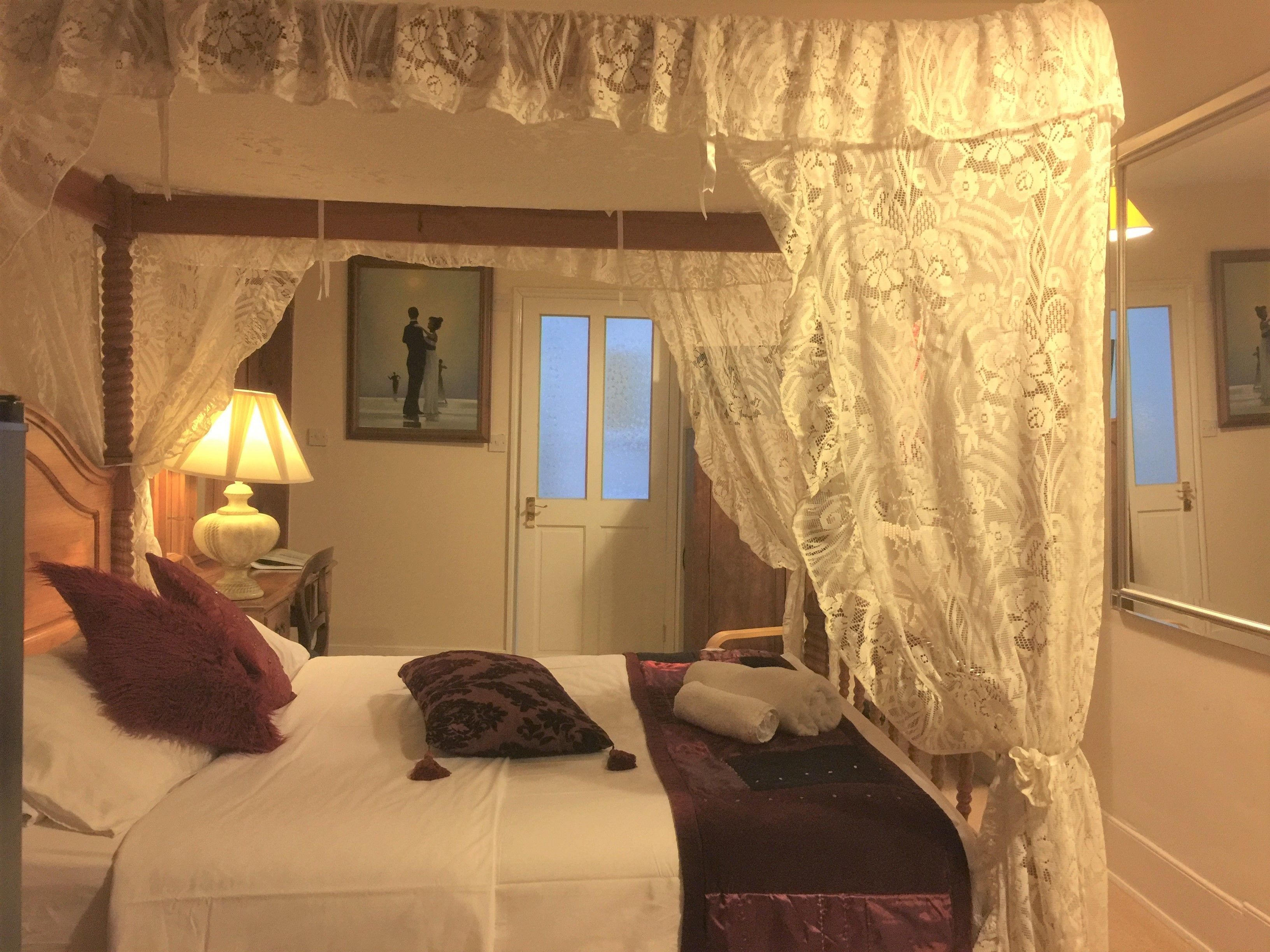 Un dormitorio con cama a dosel, una lámpara de noche, un cuadro enmarcado en la pared y una puerta con ventanas.