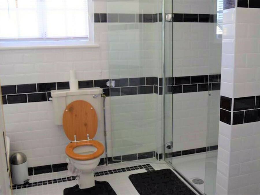 Un baño con inodoro de madera, paredes de azulejos blanco y negro, un box de ducha de vidrio y un tapete de piso con estampado en blanco y negro.
