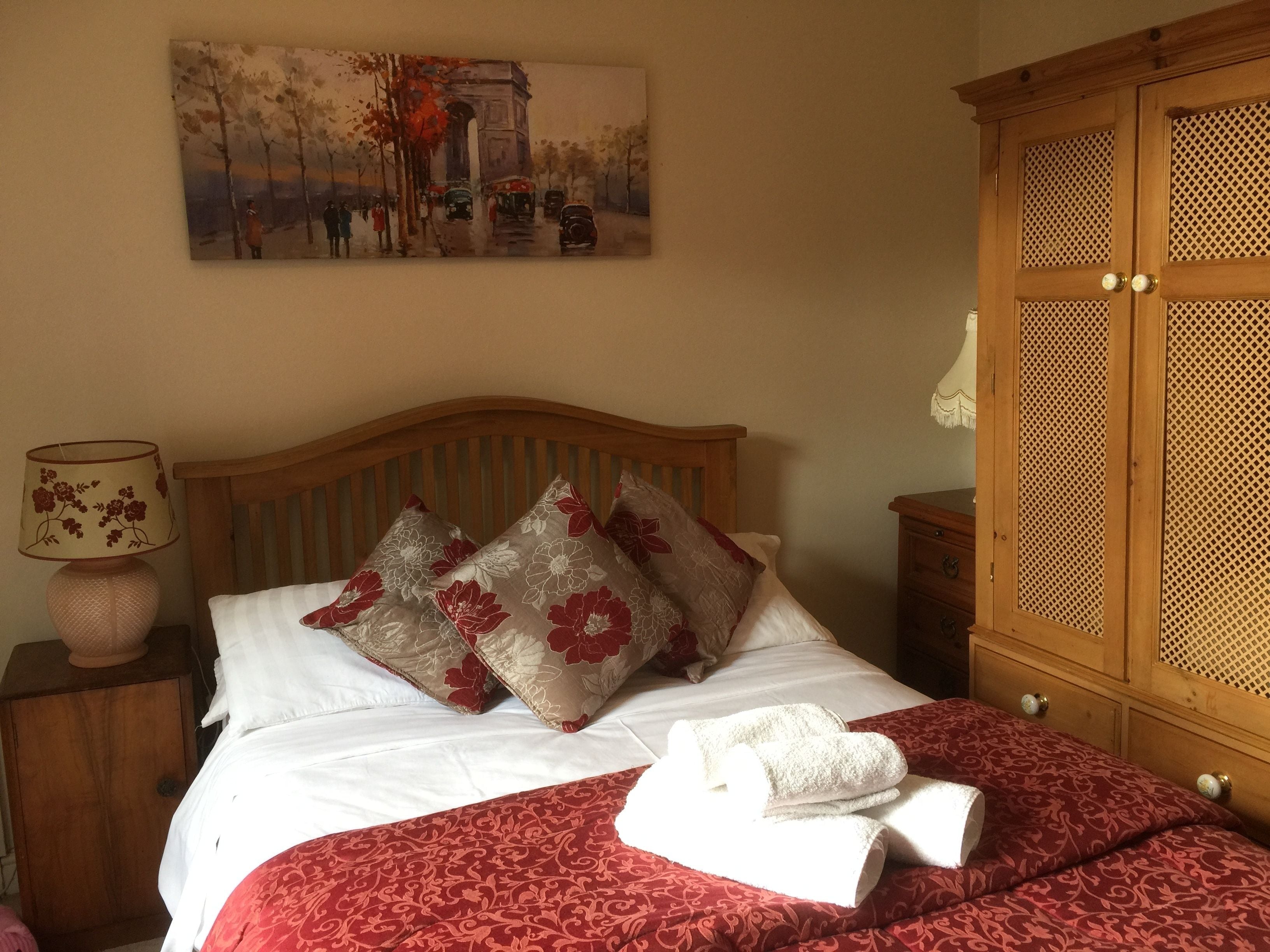 A neatly made bed with floral pillows, a red patterned bedspread, and a white towel.